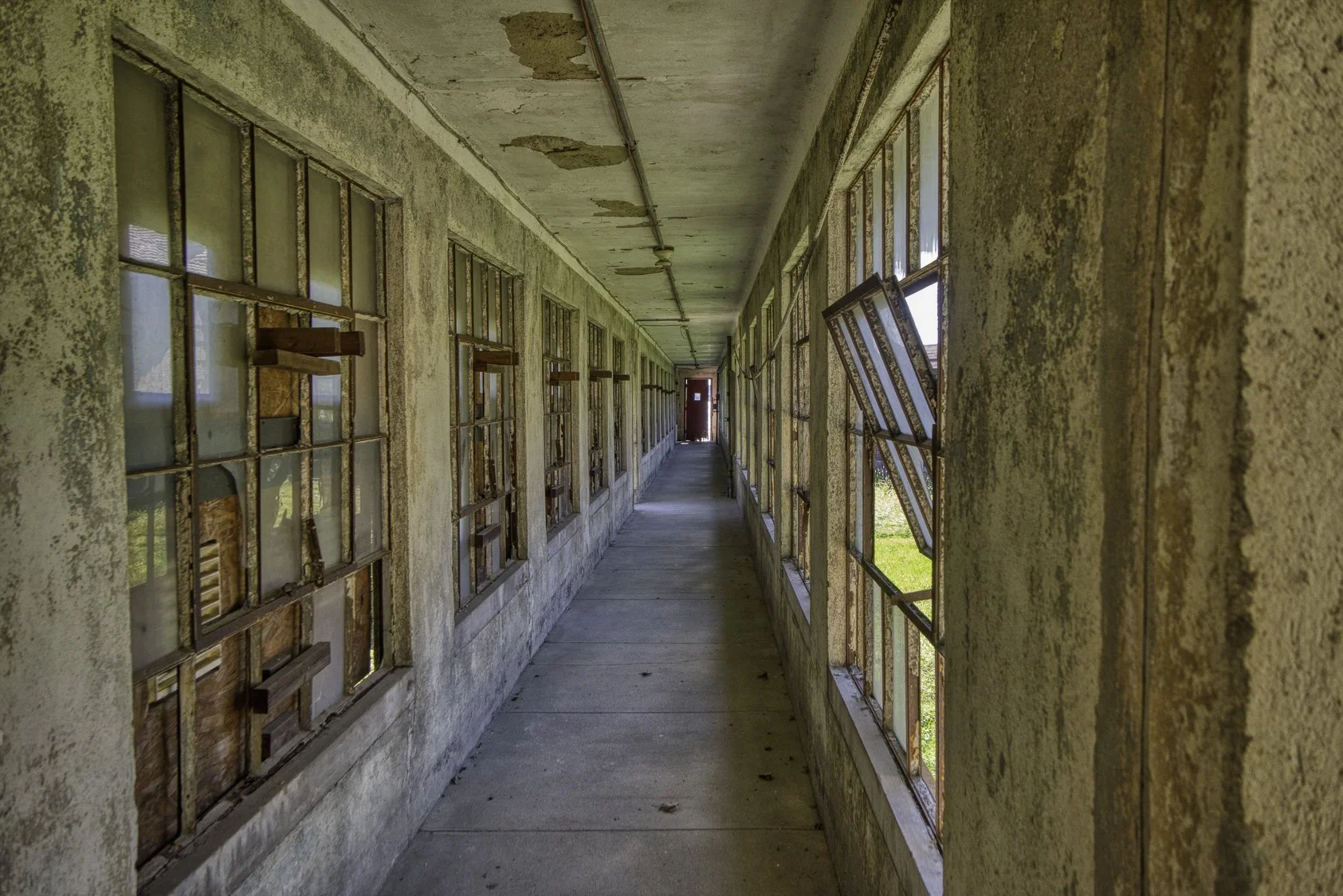 Long, abandoned hallway in a building with broken, rusty window panes and peeling walls.