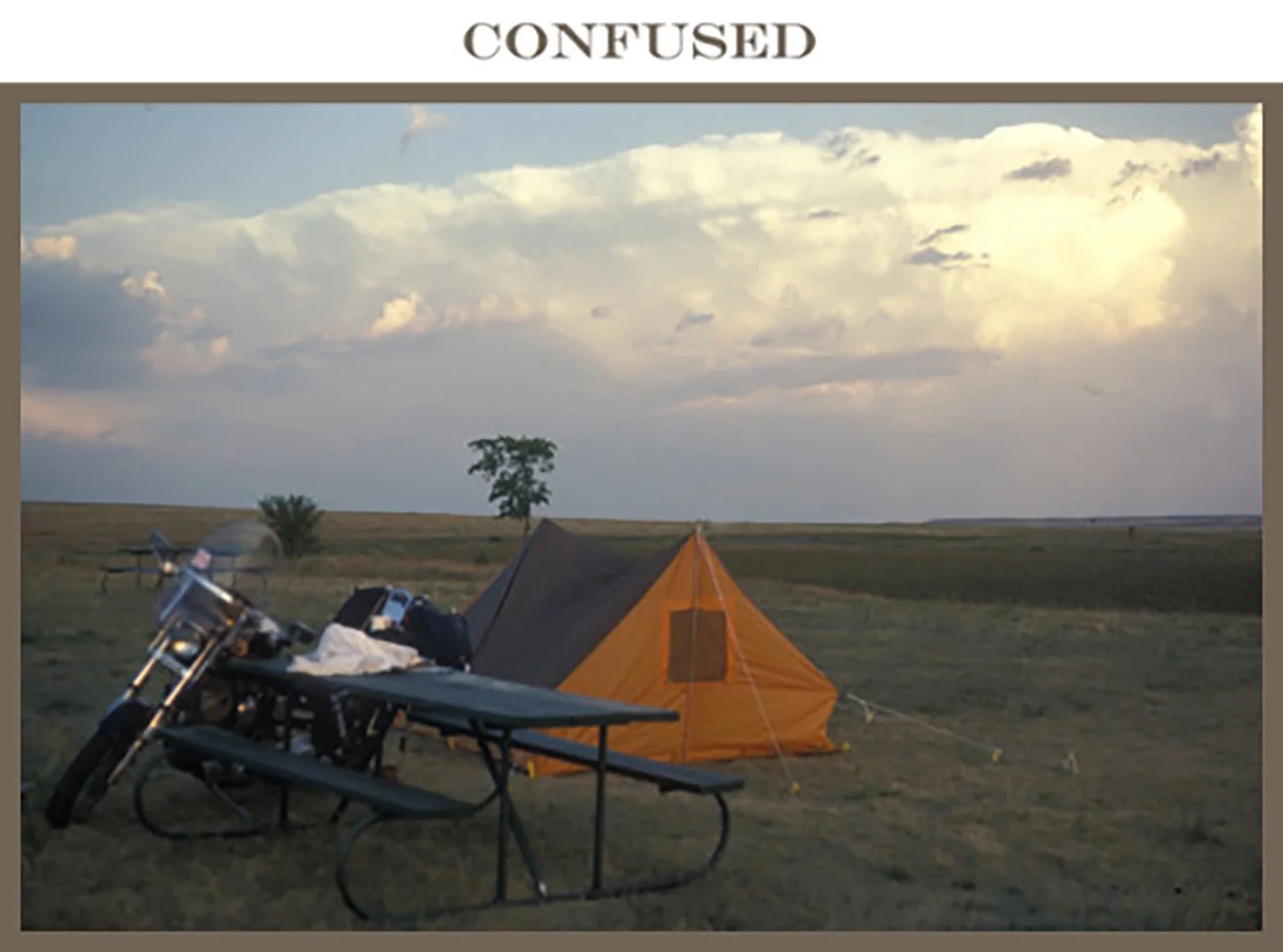 An outdoor camping scene with an orange tent, a black table, and a motorcycle on a grassy plain under a partly cloudy sky.