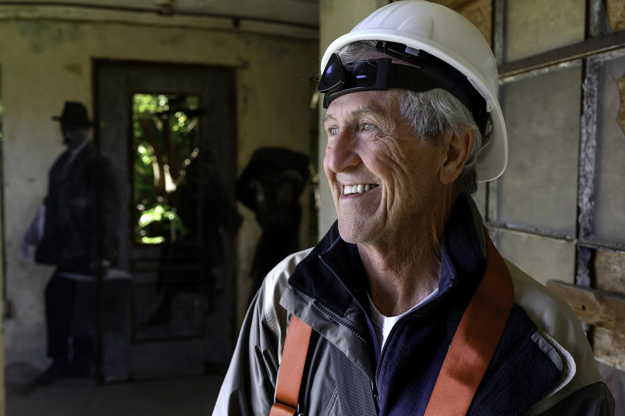 An elderly man with gray hair smiling while wearing a white safety helmet and outdoor gear, inside a rustic building with a mannequin and a mirror in the background.