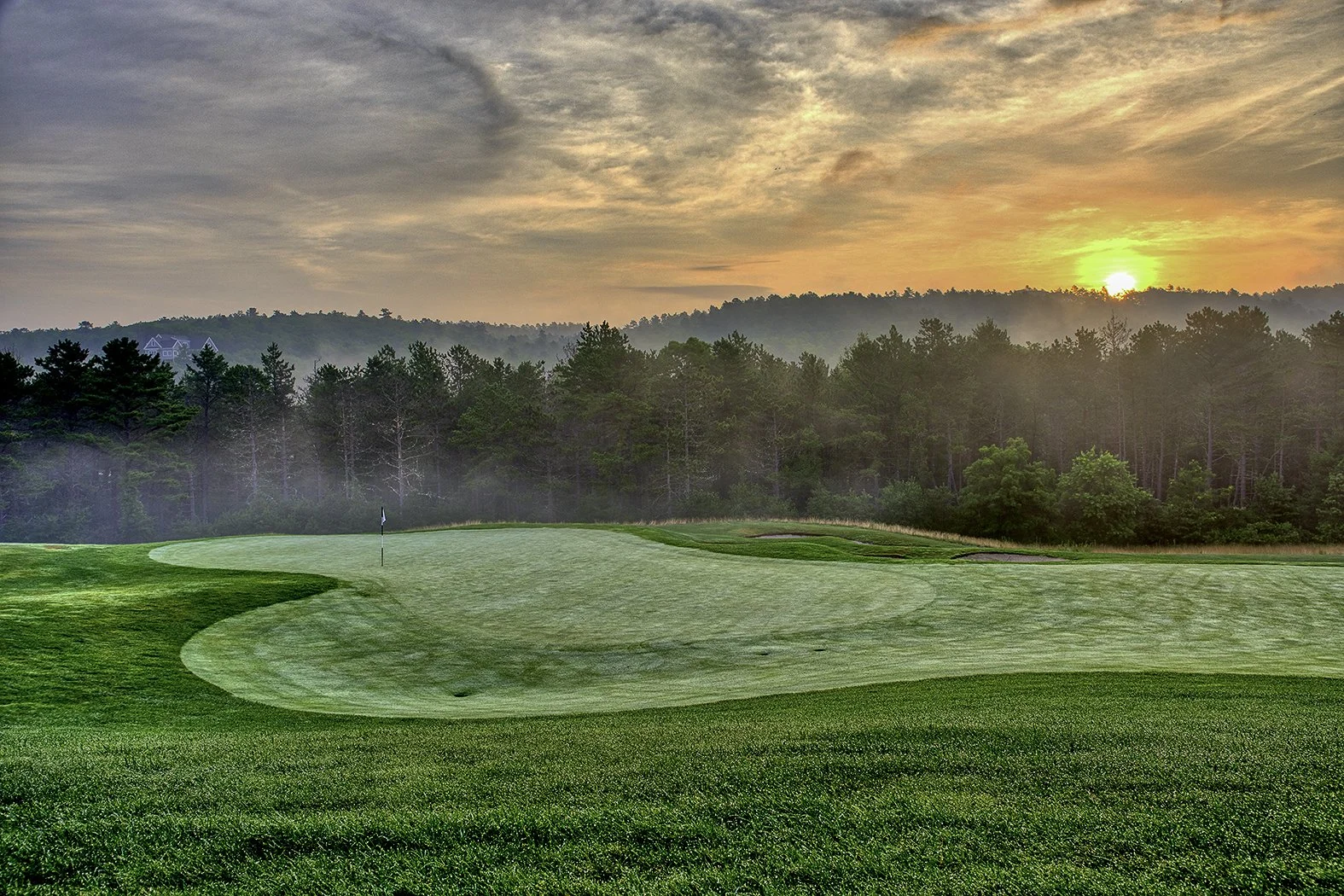 A golf course at sunset with a green, flag, and surrounding sand traps, set against a background of trees and a sunset sky.