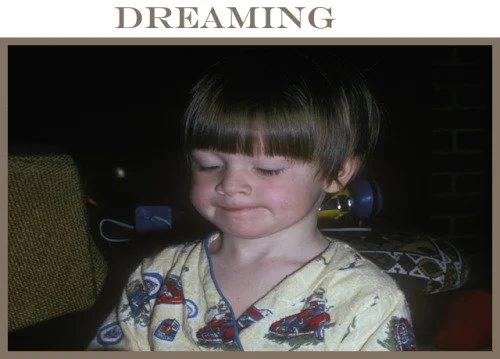 A young boy with a bowl haircut, wearing pajamas with toy cars printed on them, is smiling softly with his eyes closed.