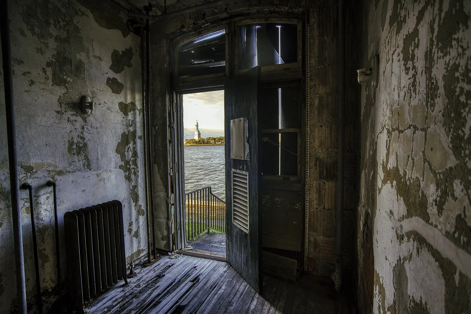 Interior of an abandoned building with peeling walls and a rusty radiator. Open door leads outside to water, with the Statue of Liberty visible in the distance across the harbor.