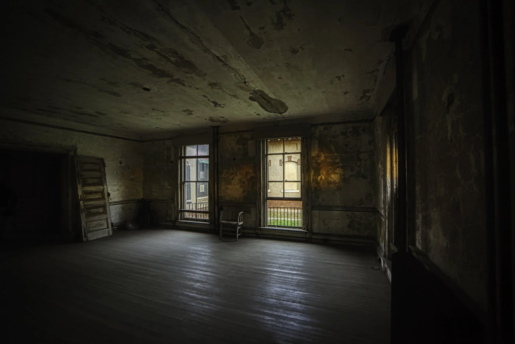 An abandoned room with peeling paint on the walls and ceiling, three windows allowing some natural light, a wooden chair, and a door leaning against the wall in the corner.