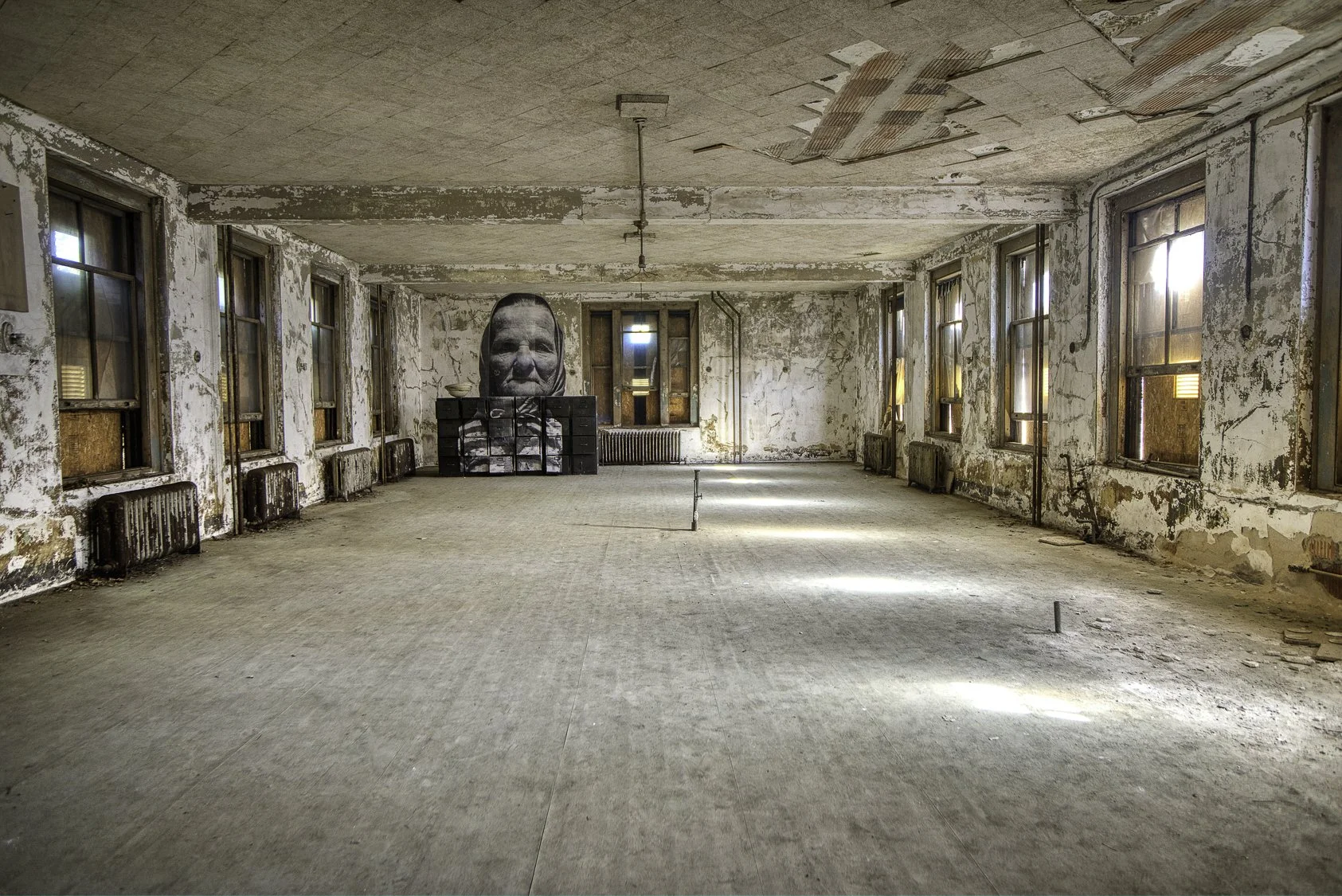 An abandoned, dilapidated room with peeling paint on walls and windows, with a large portrait of a face on black stacked boxes at the far end.