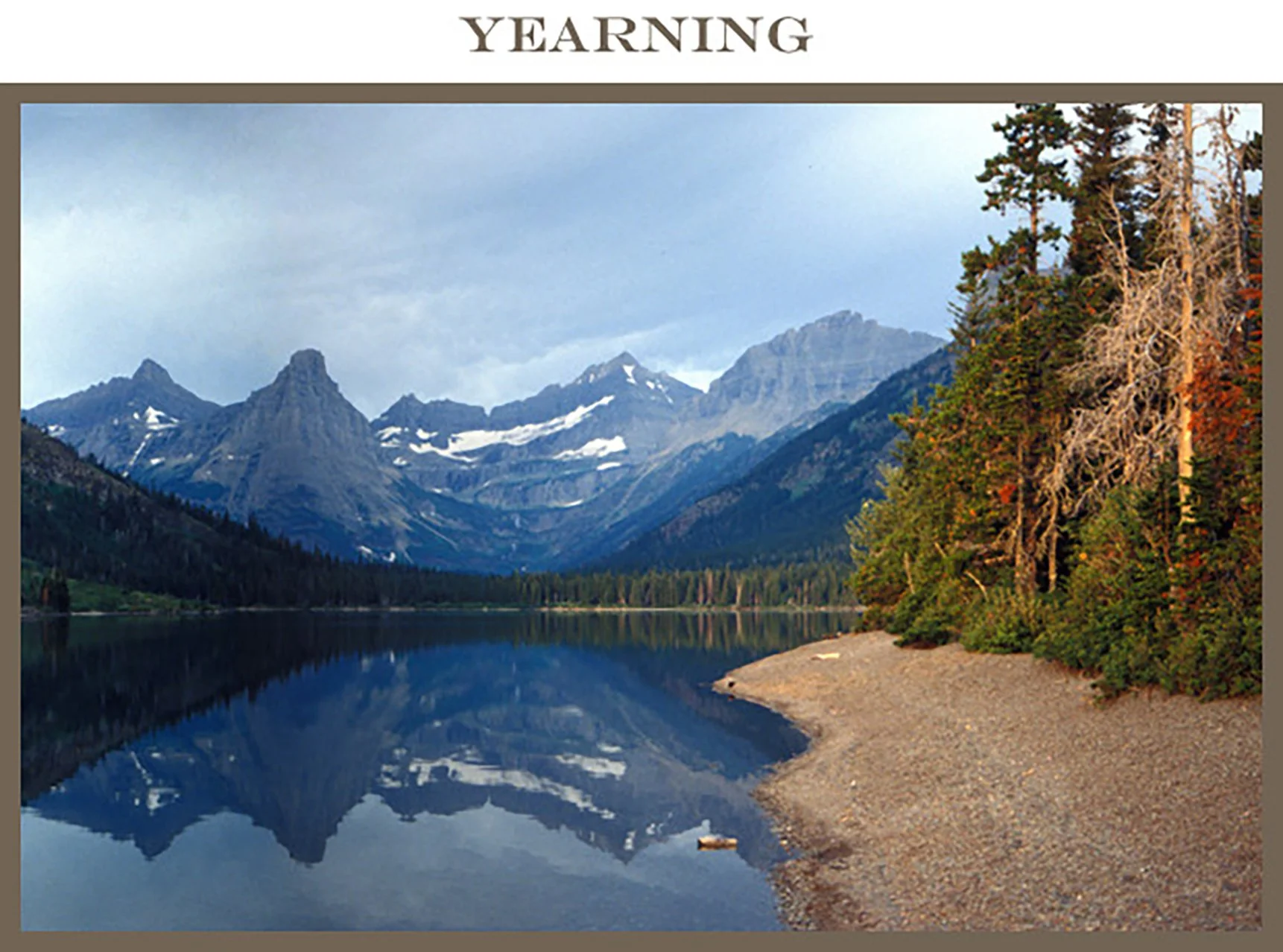 A mountain landscape with snow-capped peaks, a calm lake reflecting the mountains, and a forested shoreline with trees extending to the water's edge.