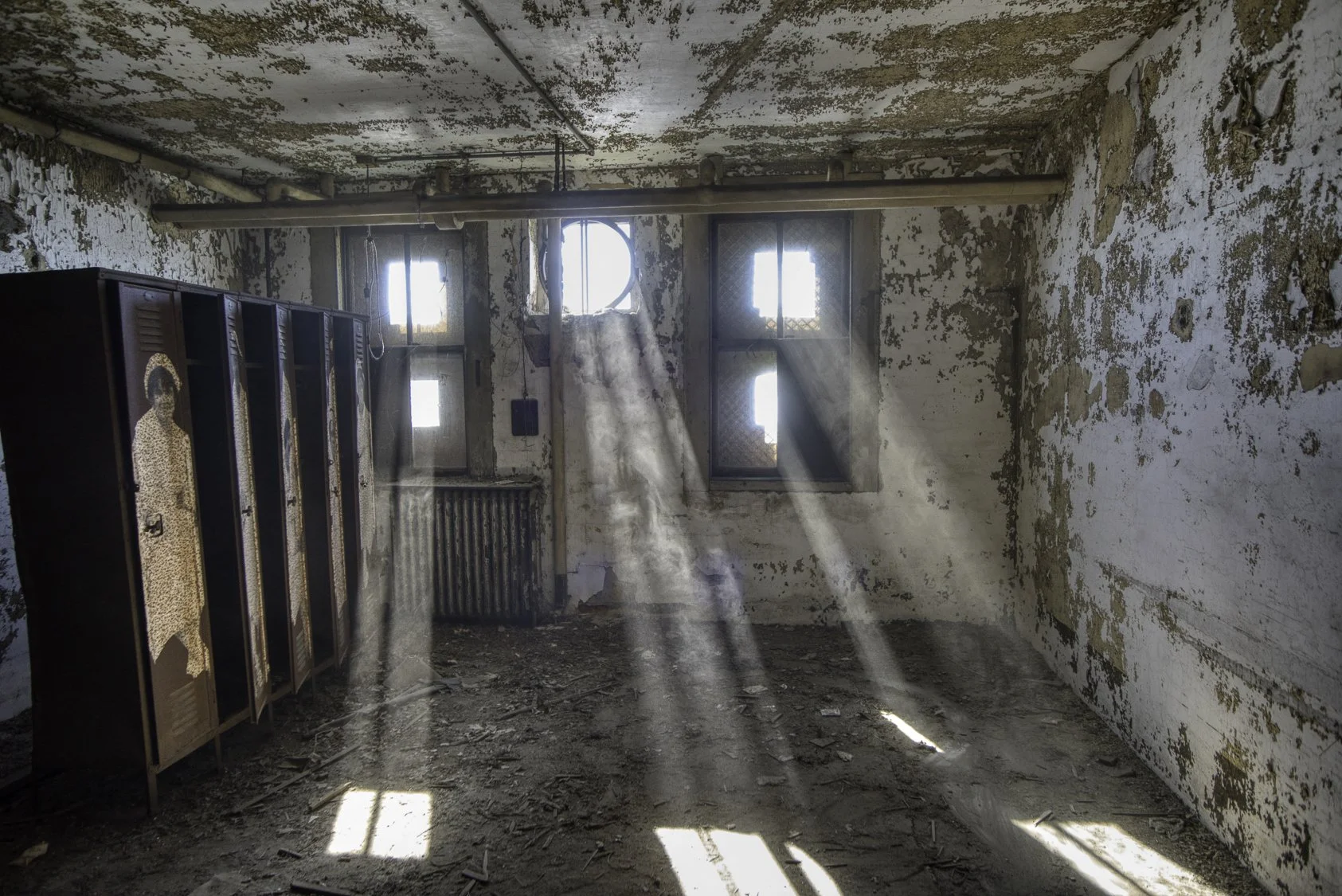 An abandoned room with peeling paint, dirt on the floor, sunlight streaming through dirty windows, and old lockers with a shuttered door.
