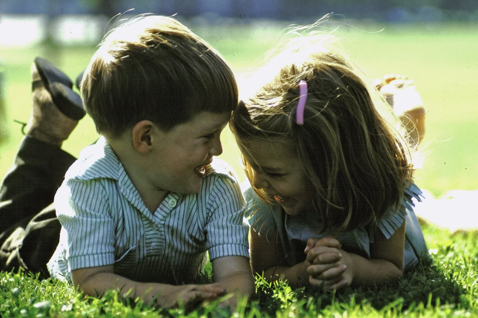 Two children, a boy and girl, are lying on the grass outdoors, smiling and laughing as they face each other with their heads close together.