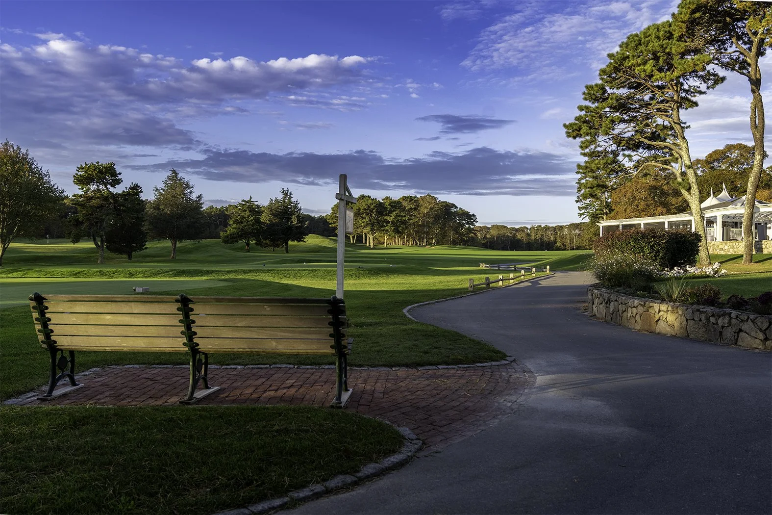 A scenic golf course with lush green grass, trees, and a blue sky with scattered clouds. There are benches along a paved pathway and a building with a white tent on the right side.