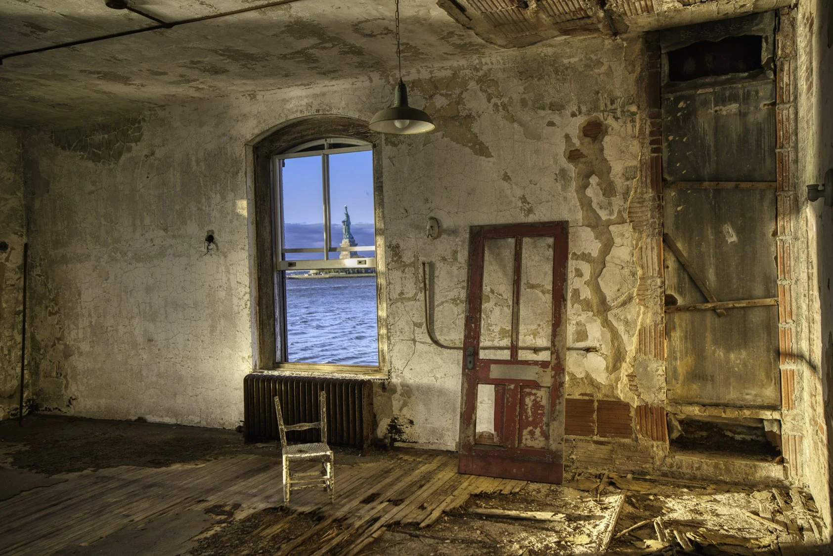 An abandoned, dilapidated room with peeling paint, broken flooring, a large window showing the Statue of Liberty across the water, an old red door leaning against the wall, and a single worn-out chair.