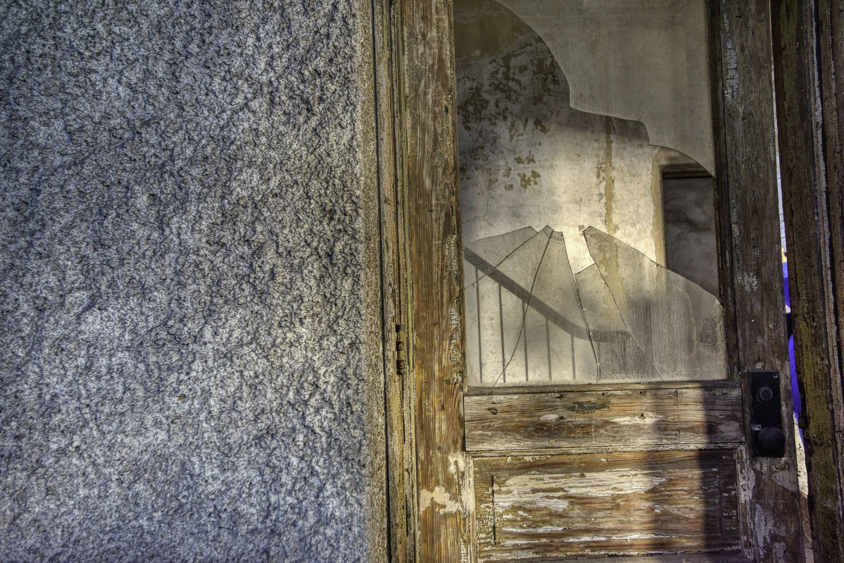 A broken glass window with a wooden frame, showing clear shards and an old, worn wall behind it.