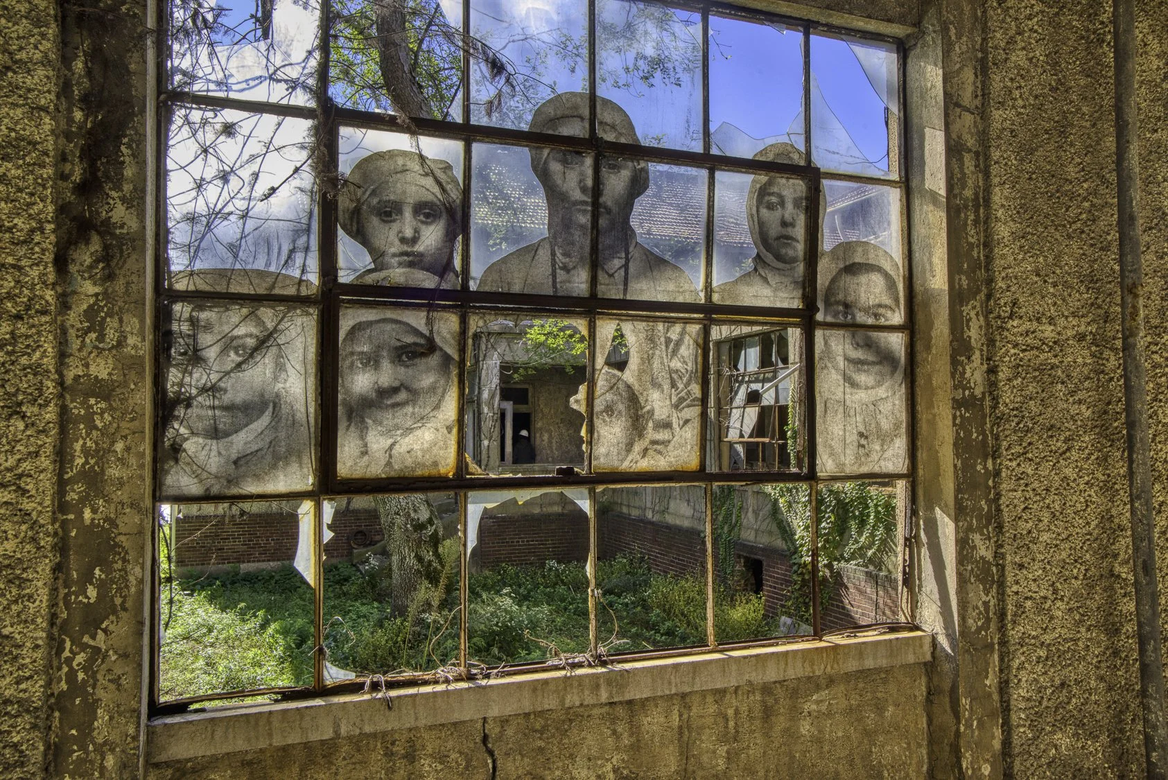 Glass window with metal grid, featuring portraits of six people taken from old photographs, with cracks and dirt on the glass, and a view of a courtyard with greenery and a building in the background.