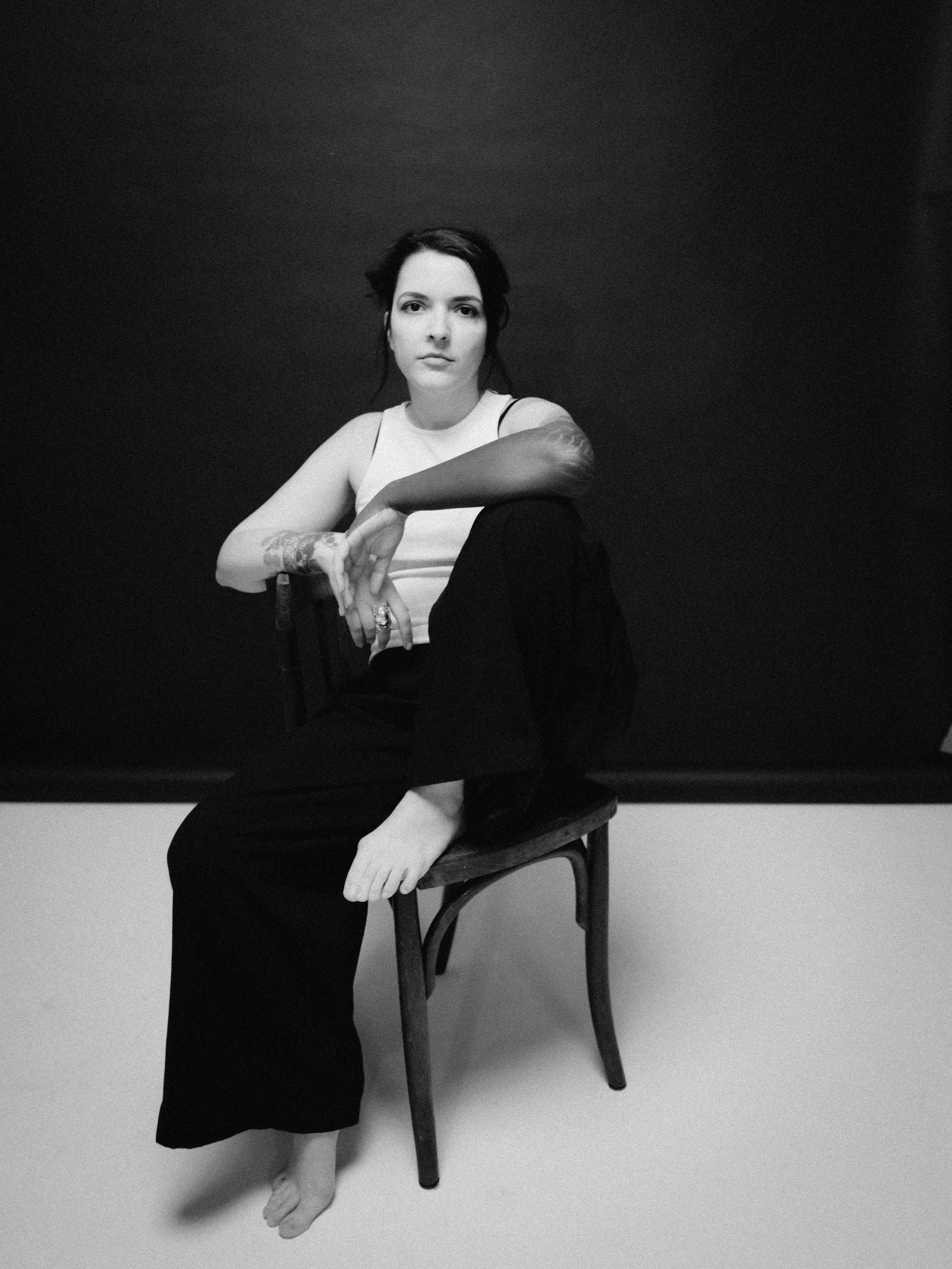 A woman sitting on a wooden chair, barefoot, with one arm resting on the chair's back, wearing a sleeveless top and long pants, against a dark background.