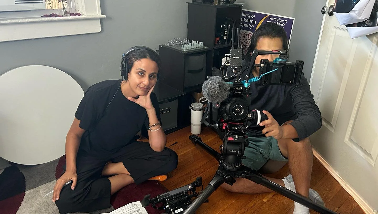 A woman and a man are filming a video indoors. The woman has dark hair, is sitting cross-legged on a carpet, wearing a black top, and has headphones on. The man is sitting on the floor behind a professional camera and rig setup, wearing grey shorts, a black long-sleeve shirt, and white shoes.