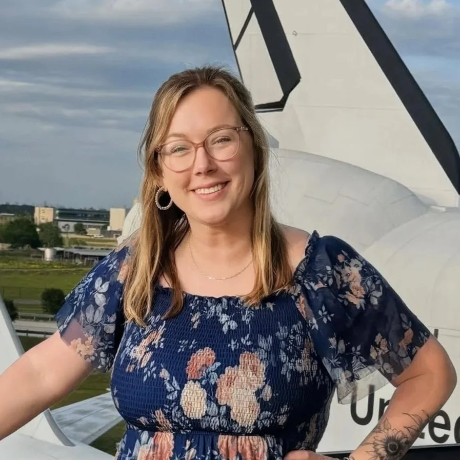 Lauren Mosis - A woman in a floral top smiles with one hand on her hip in front of an aircraft 