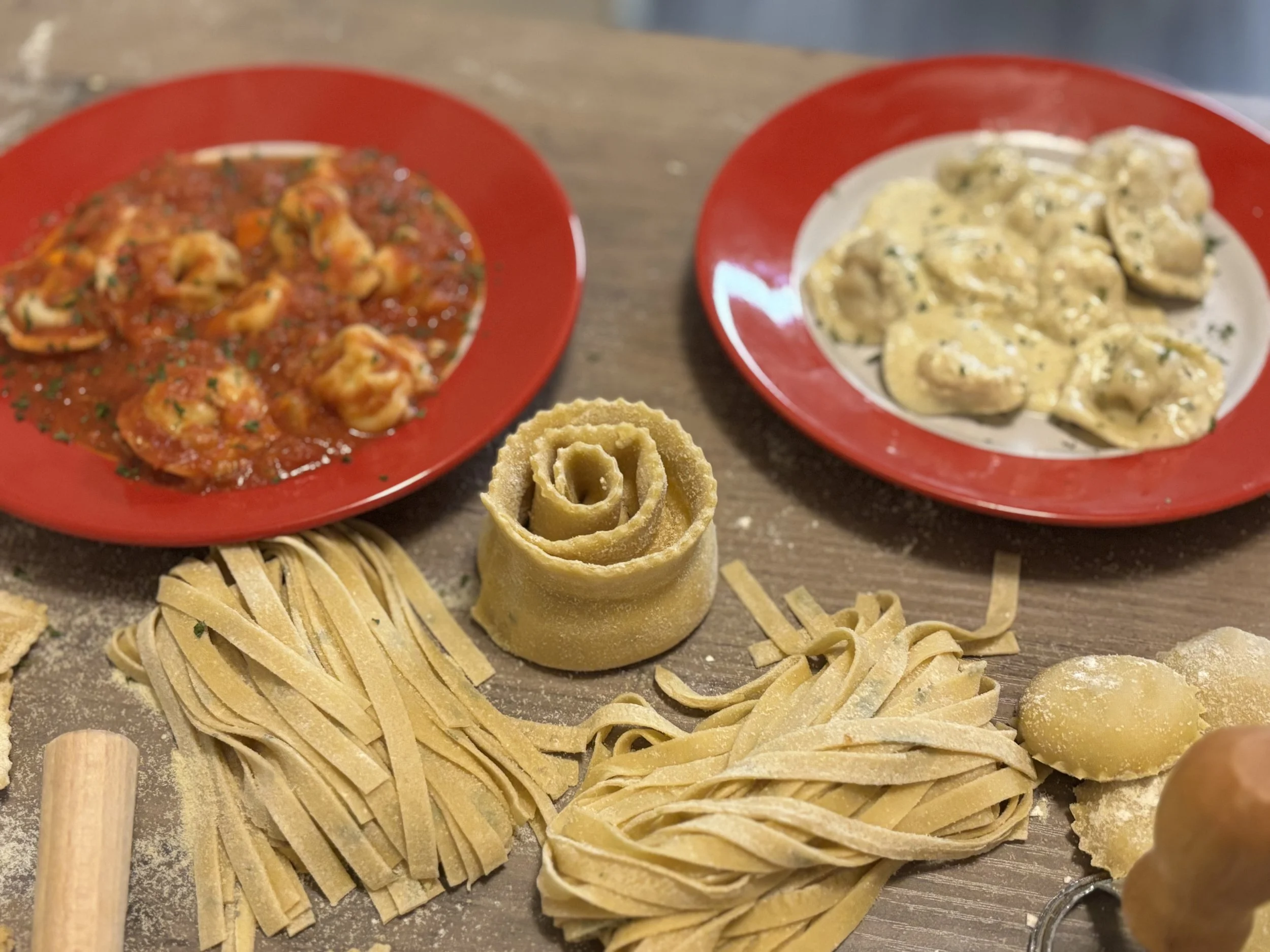 Various types of pasta and two plates of sauces on a wooden surface.