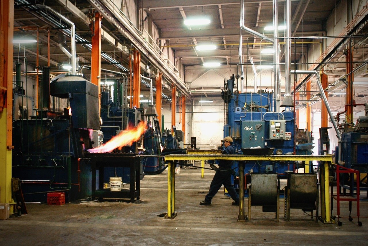 A worker is on a factory floor pulling machine parts of a large, industrial oven. A large flame spits out of the oven as he pulls the metal parts out on a rack