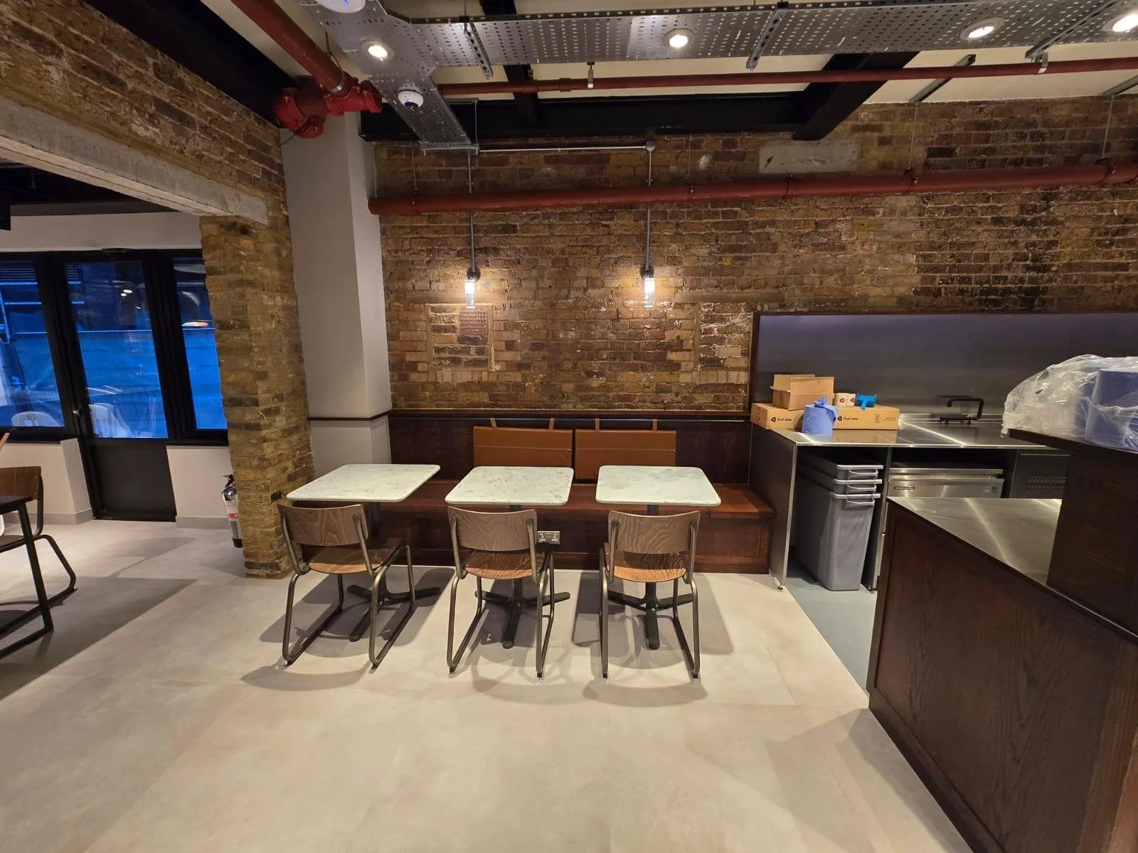 Interior of a restaurant with brick walls, three tables with chairs, and a kitchen area with boxes and appliances.