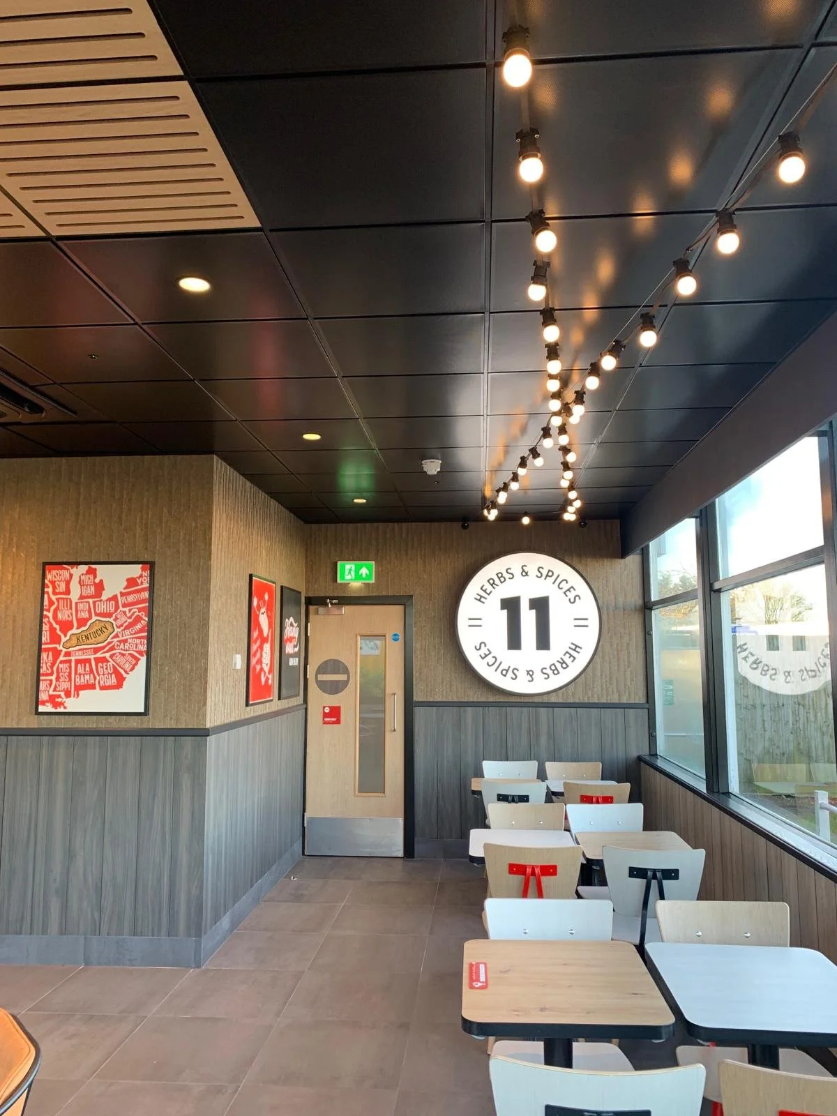 Interior of a fast-food or casual dining restaurant. There are several white and light wood chairs arranged around tables. A large round sign on the wall reads 'Herbs & Spices 11'. The wall has framed pictures, and there is a door with a no-entry sign. Large windows on the right let in natural light.