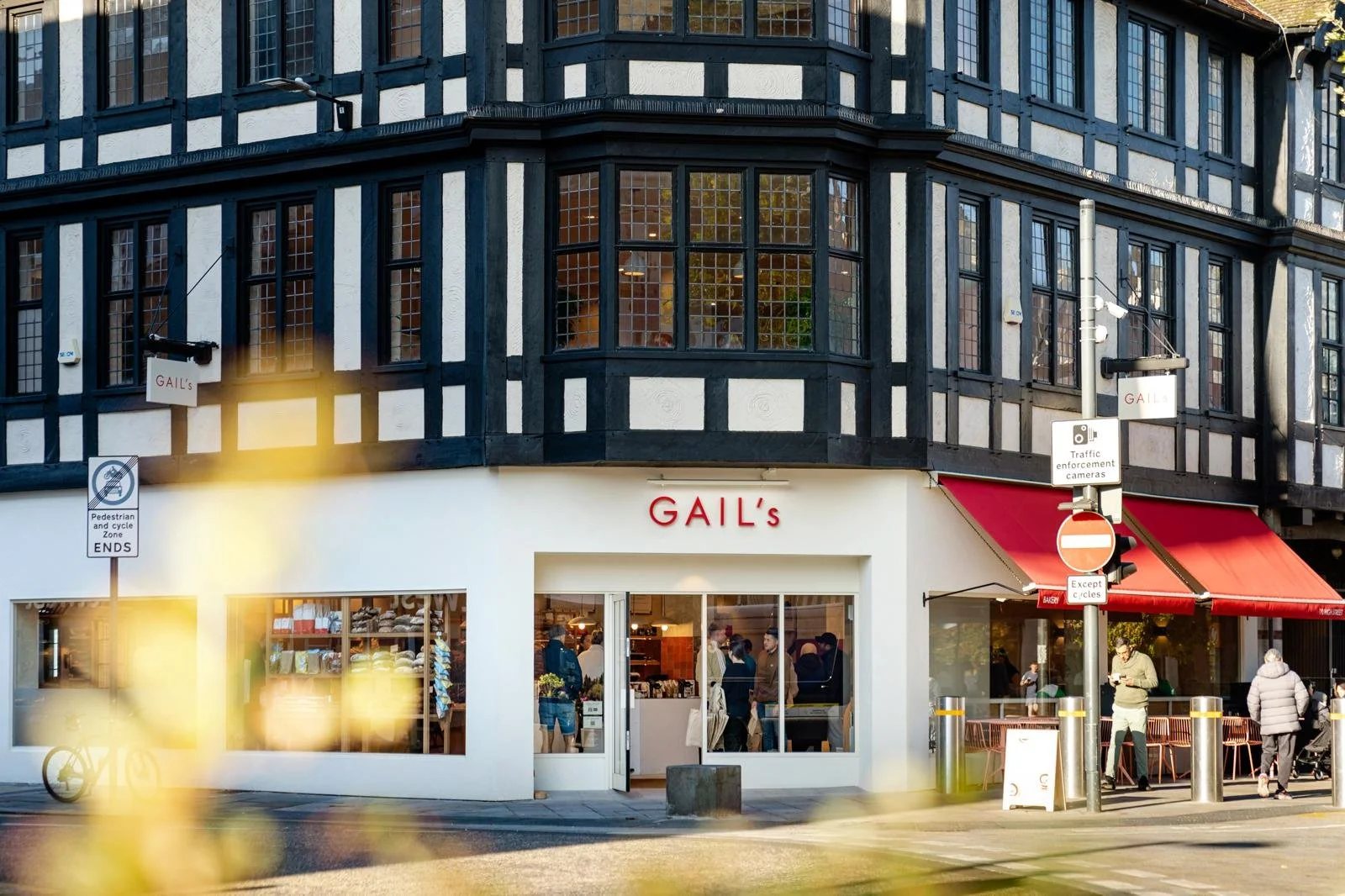 Street view of a storefront named GAIL's with large windows and a red awning, people inside and outside, traffic signs, and a corner building with black and white timber framing.