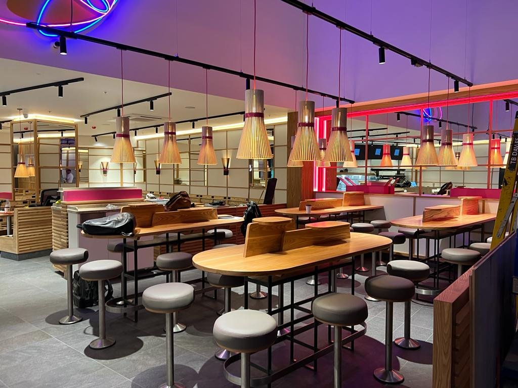 Empty modern restaurant interior with wooden tables, stool seating, and hanging pendant lights with neon lighting accents.
