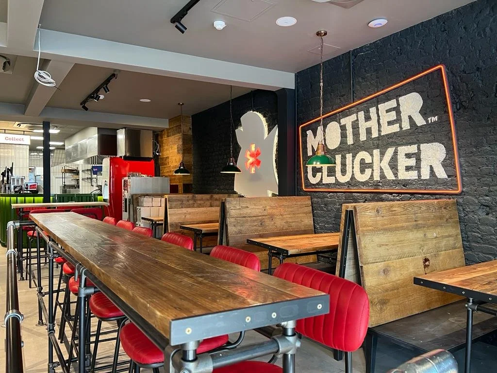 Interior of a restaurant with a long wooden table and red chairs, black brick wall with a neon sign reading 'Mother Clucker', and wooden booths.