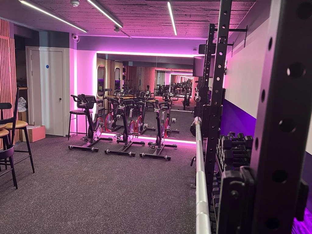 An indoor gym with exercise bikes, a mirror wall, and weightlifting equipment, illuminated with pink neon lighting.