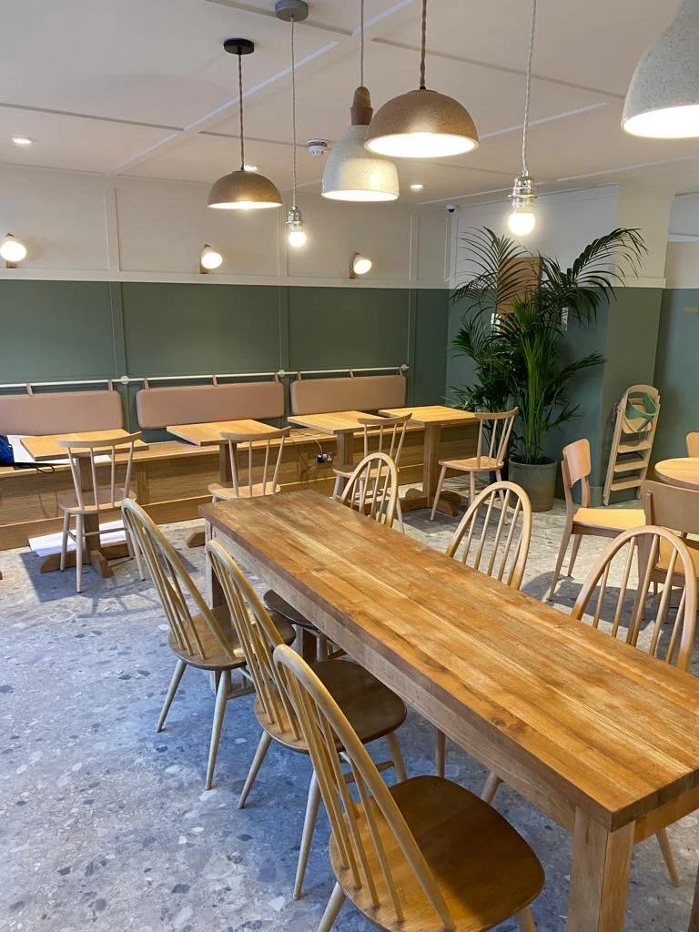 Empty cafe or restaurant with wooden tables and chairs, pendant lights hanging from ceiling, green walls, and a large potted plant.