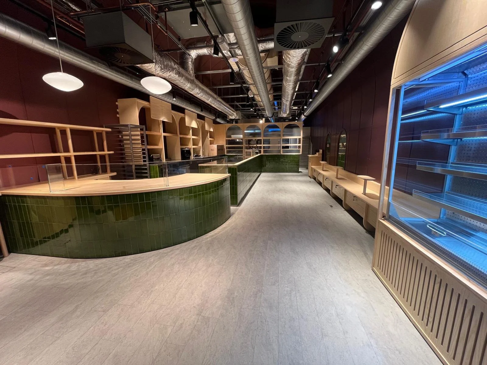 Empty retail or food store interior with green tiled counter, wooden shelves, and an empty refrigerated display case on the right, with overhead industrial ductwork and lighting.
