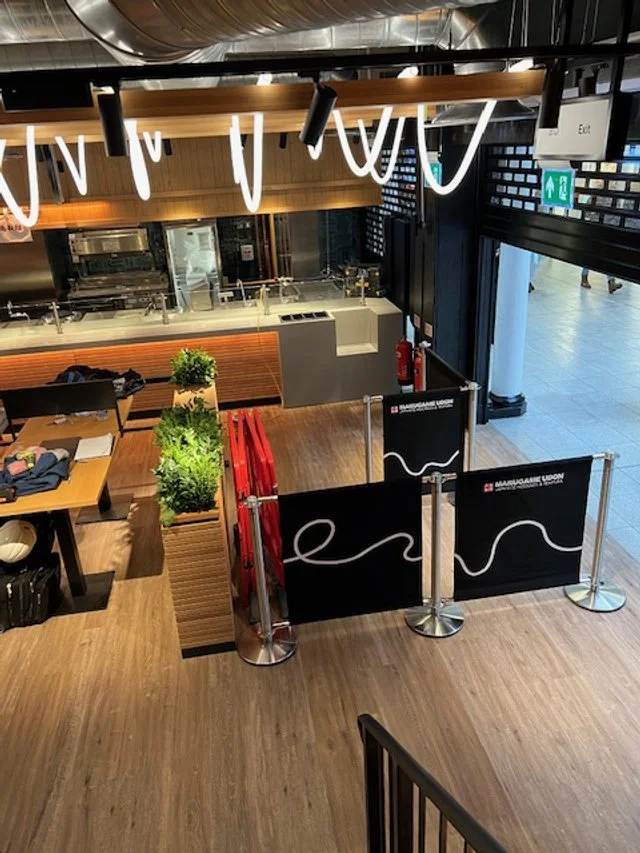 Interior view of a restaurant or food court with a counter, tables, and plants, seen from an upper angle.