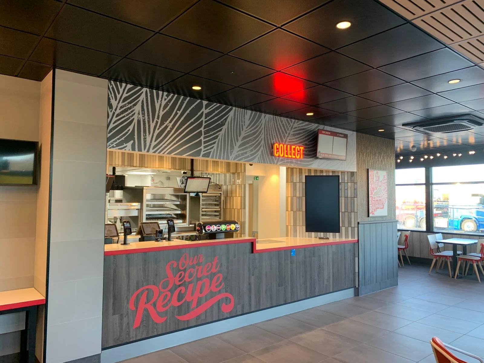 Empty fast food restaurant counter with a sign that reads 'Our Secret Recipe' and a red neon sign that says 'Collect' inside a modern dining area with seating and large windows.