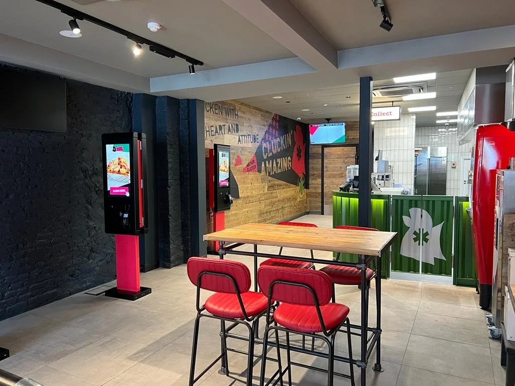 Inside a fast-food restaurant with two self-order kiosks, a wooden table with three red chairs, a counter area with a green panel, a red soda vending machine, and a mural with colorful text on the wall.