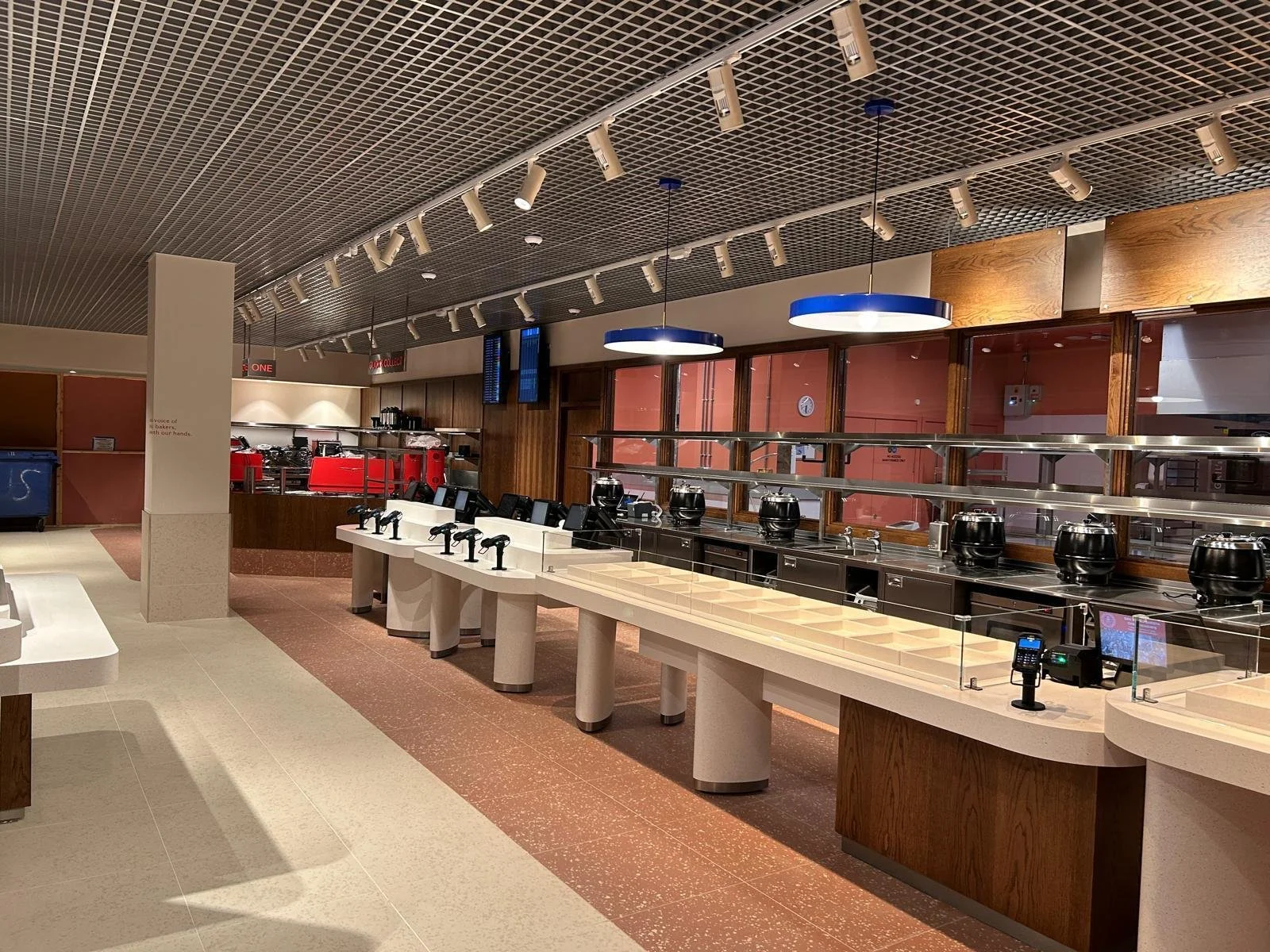 Empty cafeteria serving area with conveyor belt, utensils, and warming pots, illuminated by round ceiling lights, with wooden and glass decor.
