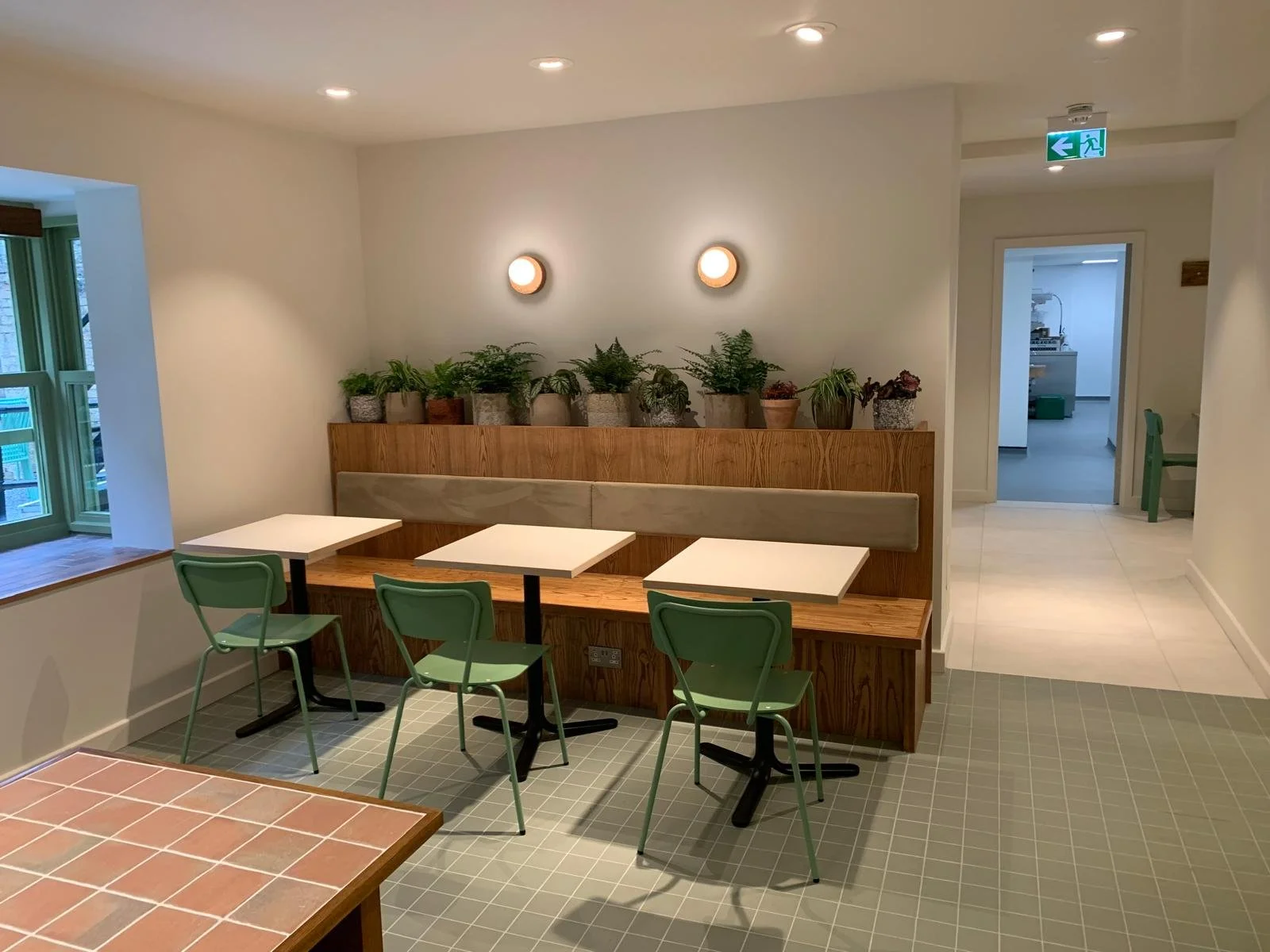 Interior of a cozy cafe or dining area with green chairs, white tables, potted plants on a wooden ledge, and soft wall-mounted lights.