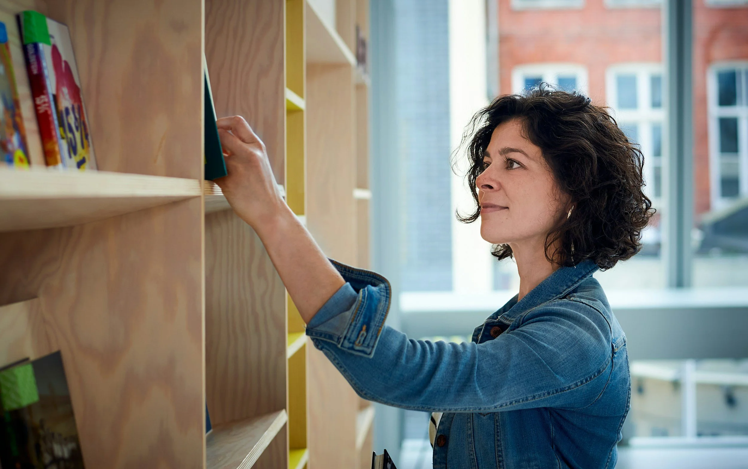 A woman with dark curly hair wearing a denim jacket puts a book on a wooden bookshelf in a bright room with large windows.