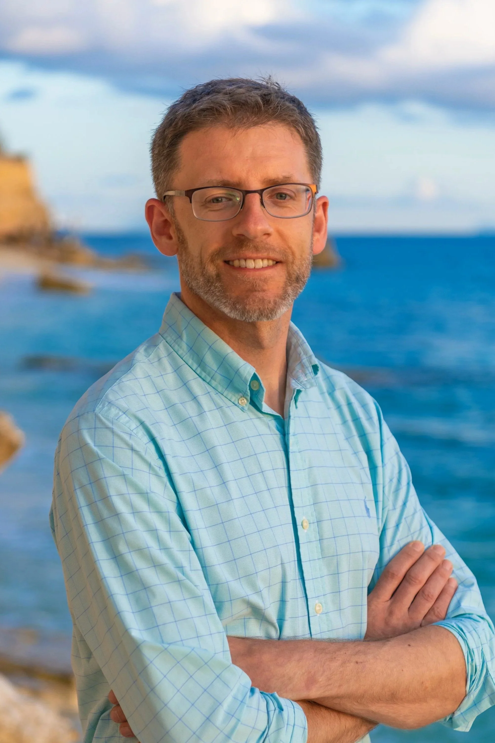 A man with glasses and a beard smiling at the camera, standing by the ocean with cloudy sky and cliffs in the background.
