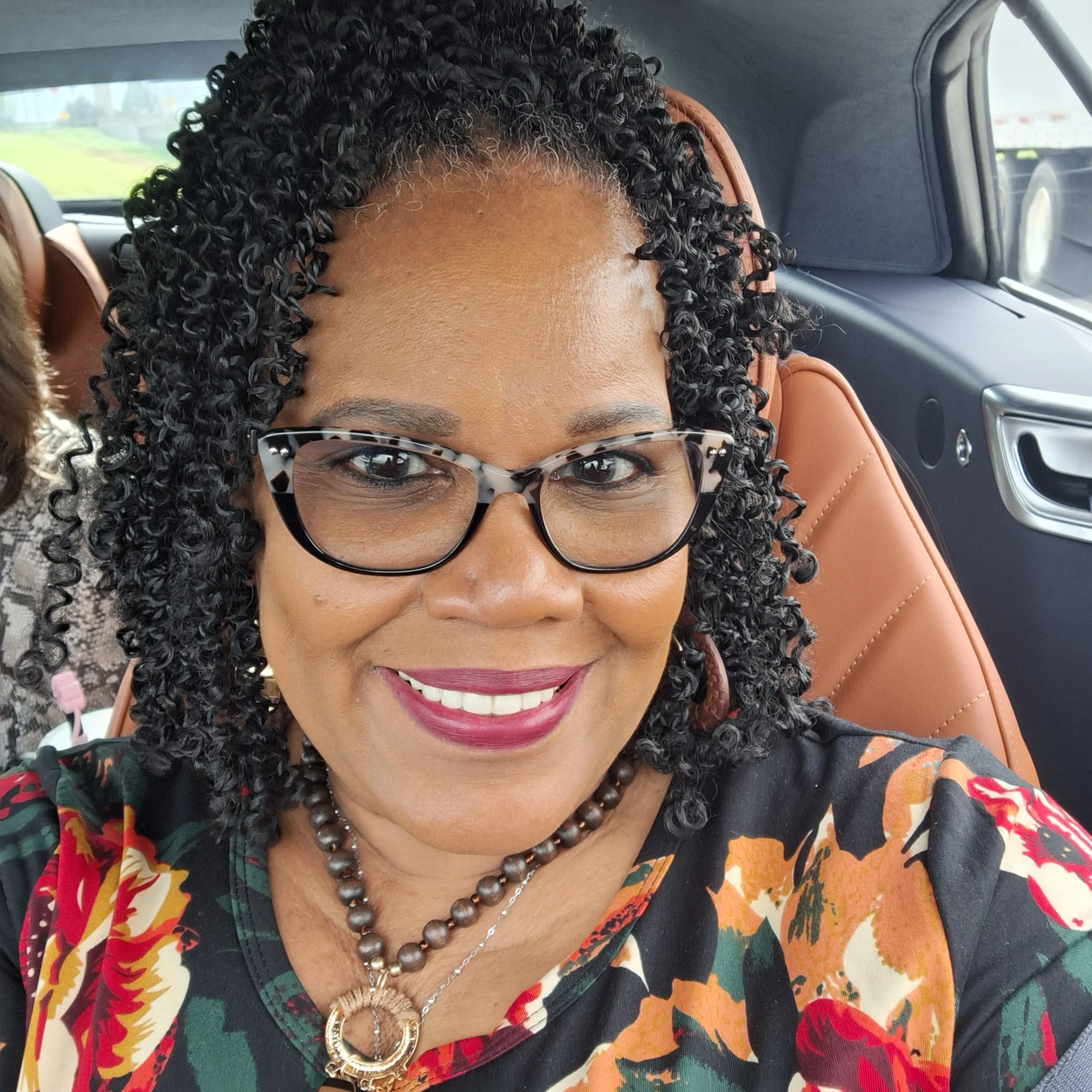 A woman with curly black hair, glasses, and lipstick, smiling for a selfie inside a vehicle with brown leather seats, wearing a patterned blouse and a beaded necklace.