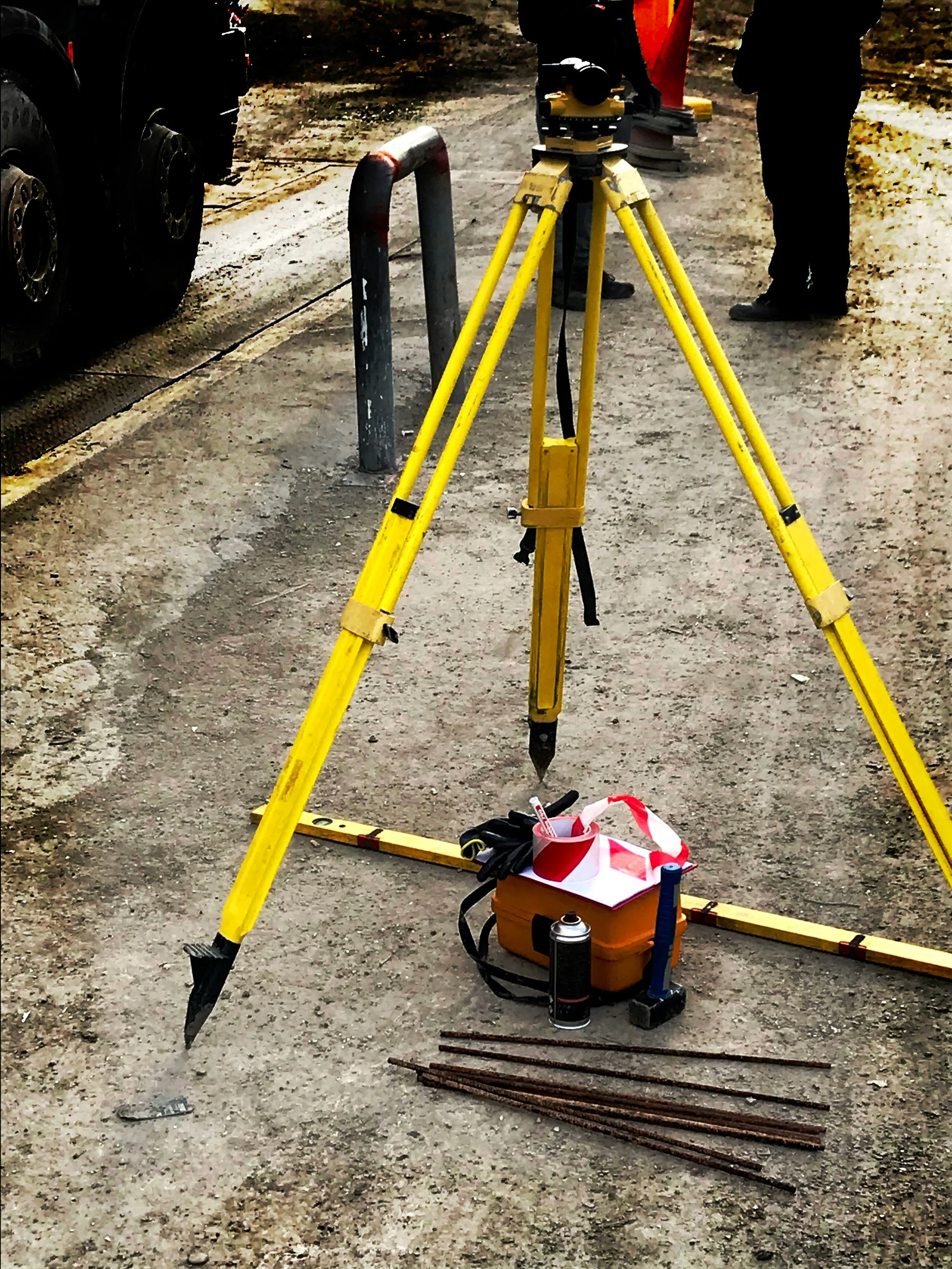 Surveying equipment on a construction site, including a yellow tripod with a rotating device on top, resting on a dirt surface along with some tools and supplies.