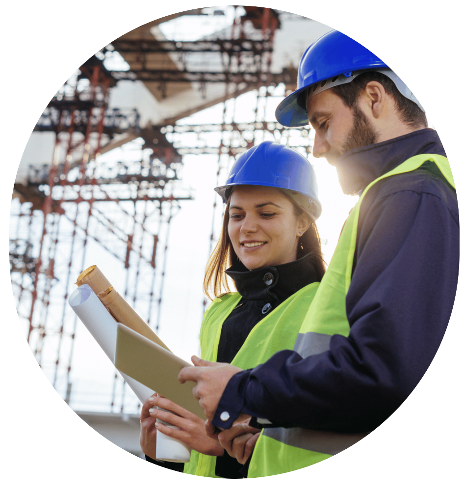 Two construction workers, a woman and a man, wearing blue hard hats and high-visibility vests, standing outdoors at a construction site, looking at blueprints and smiling.