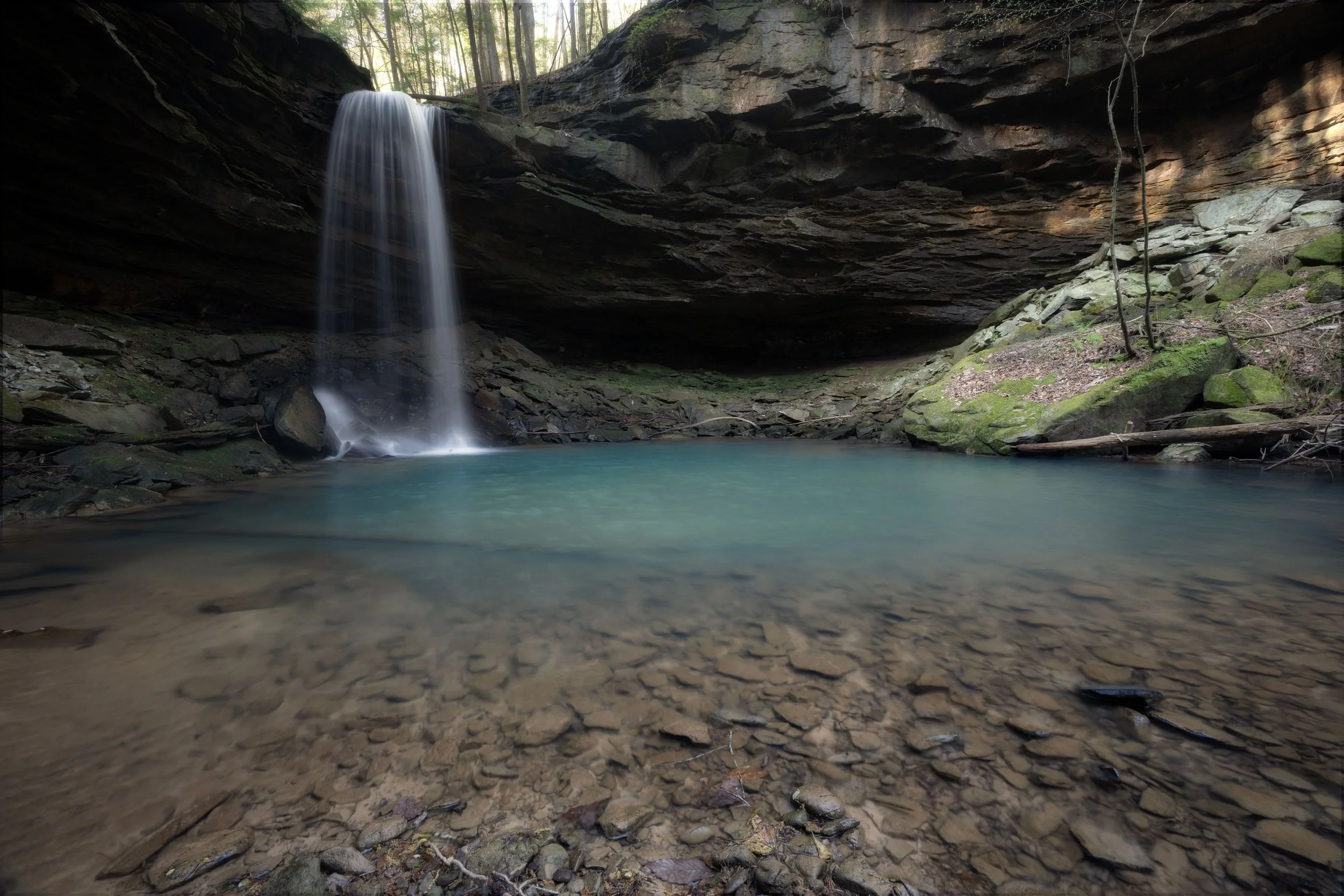 A small waterfall cascading into a turquoise pool in a rocky canyon, surrounded by trees and moss-covered rocks.