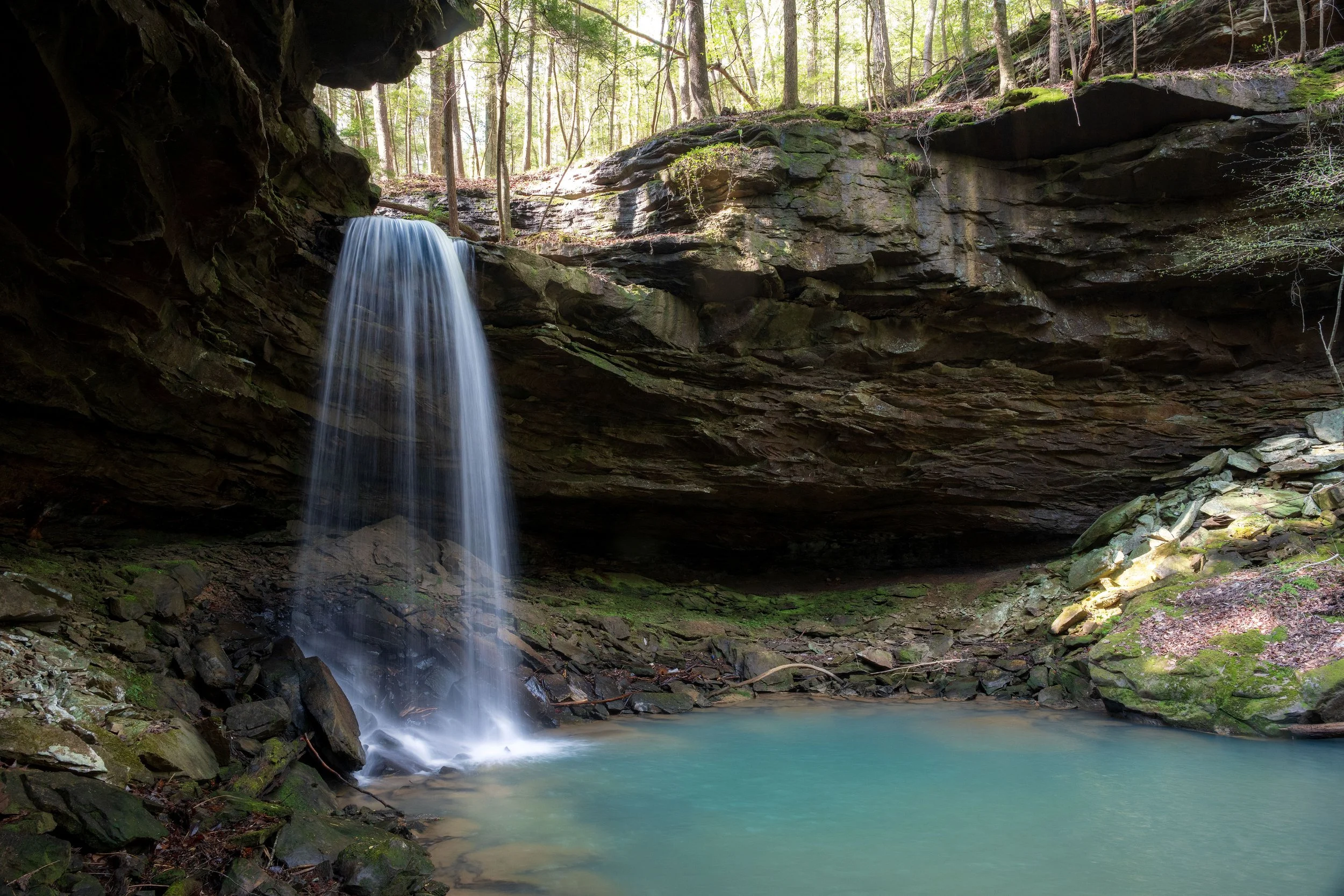 A waterfall cascading into a turquoise pool in a forest surrounded by rocks and trees, with sunlight filtering through the leaves.