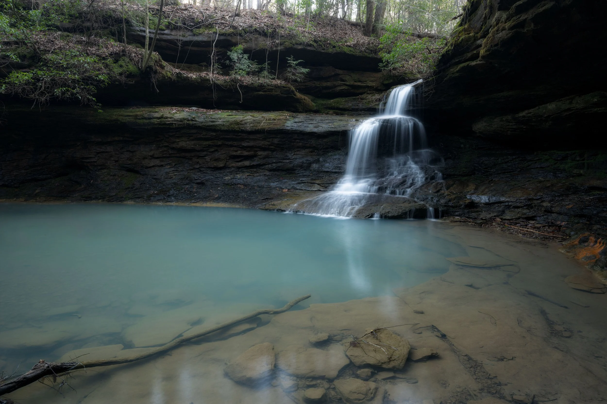 A small waterfall cascading into a clear, shallow pool in a wooded area.