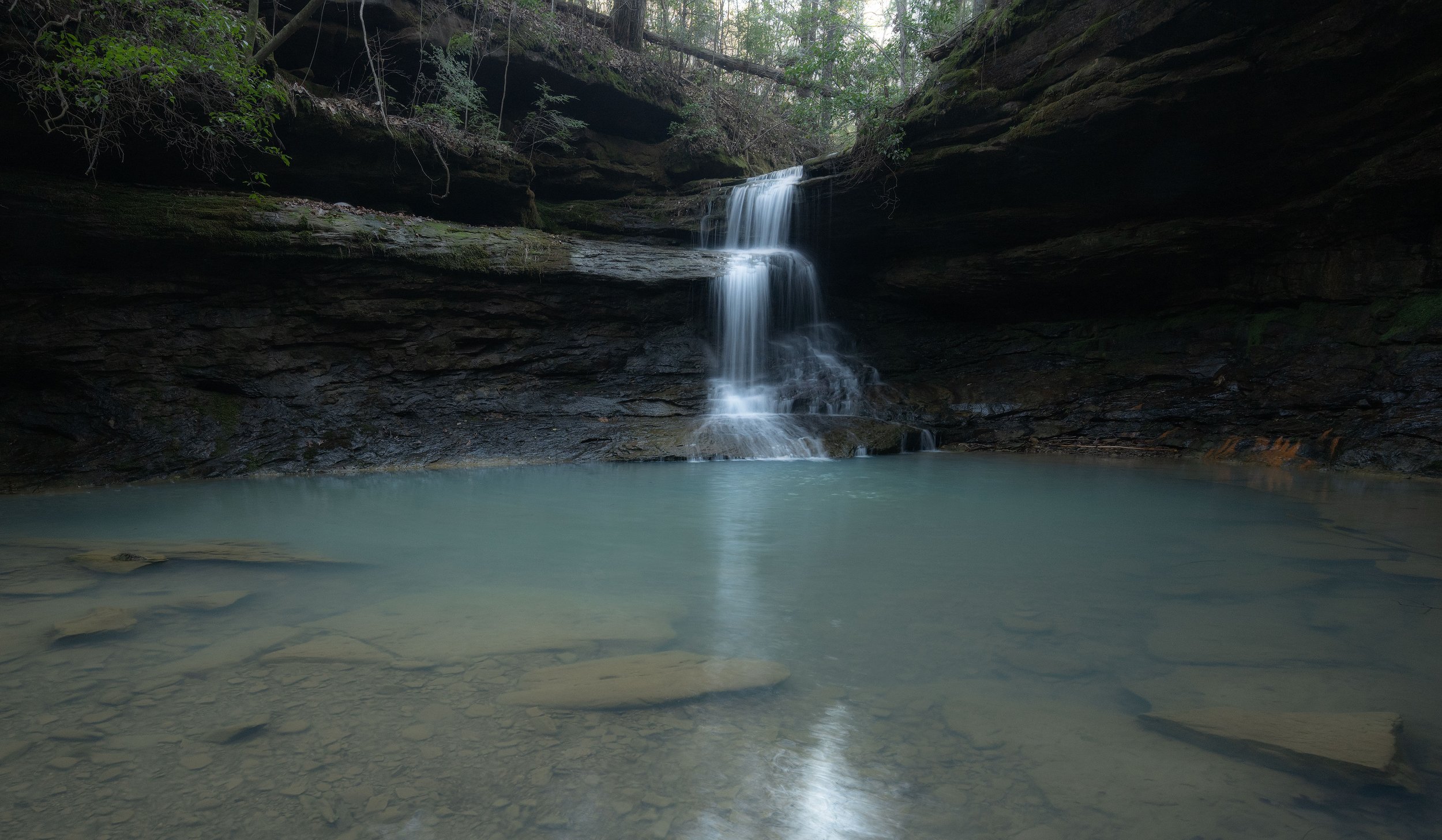 Flowing waterfall over rocky ledge into a serene pool of water in a forested area.