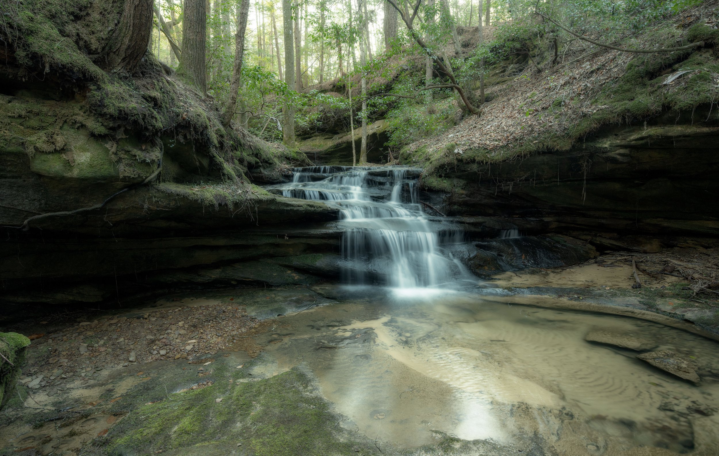 Waterfall cascading over rocks in a lush, green forest with sunlight filtering through the trees.