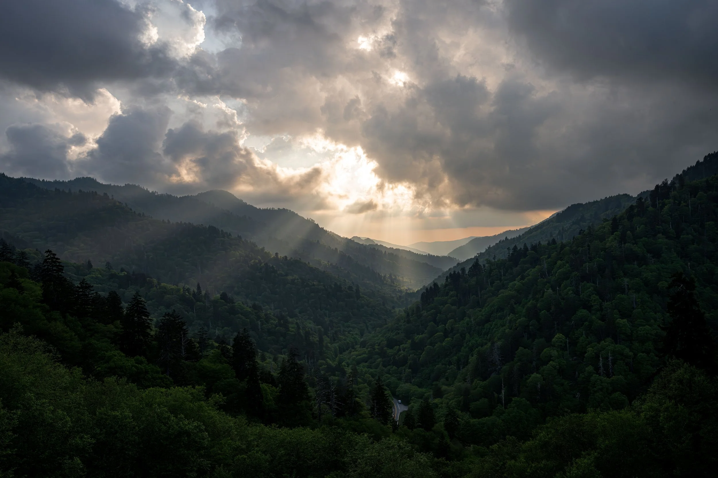 Sunlight breaking through dark clouds over a green mountain valley with dense forest and distant mountain ridges.
