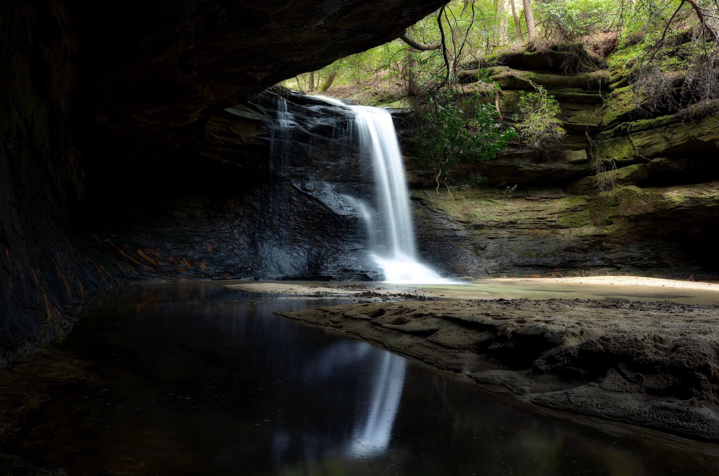 A small waterfall flowing into a calm pool of water inside a dark, rocky cave with some greenery outside.
