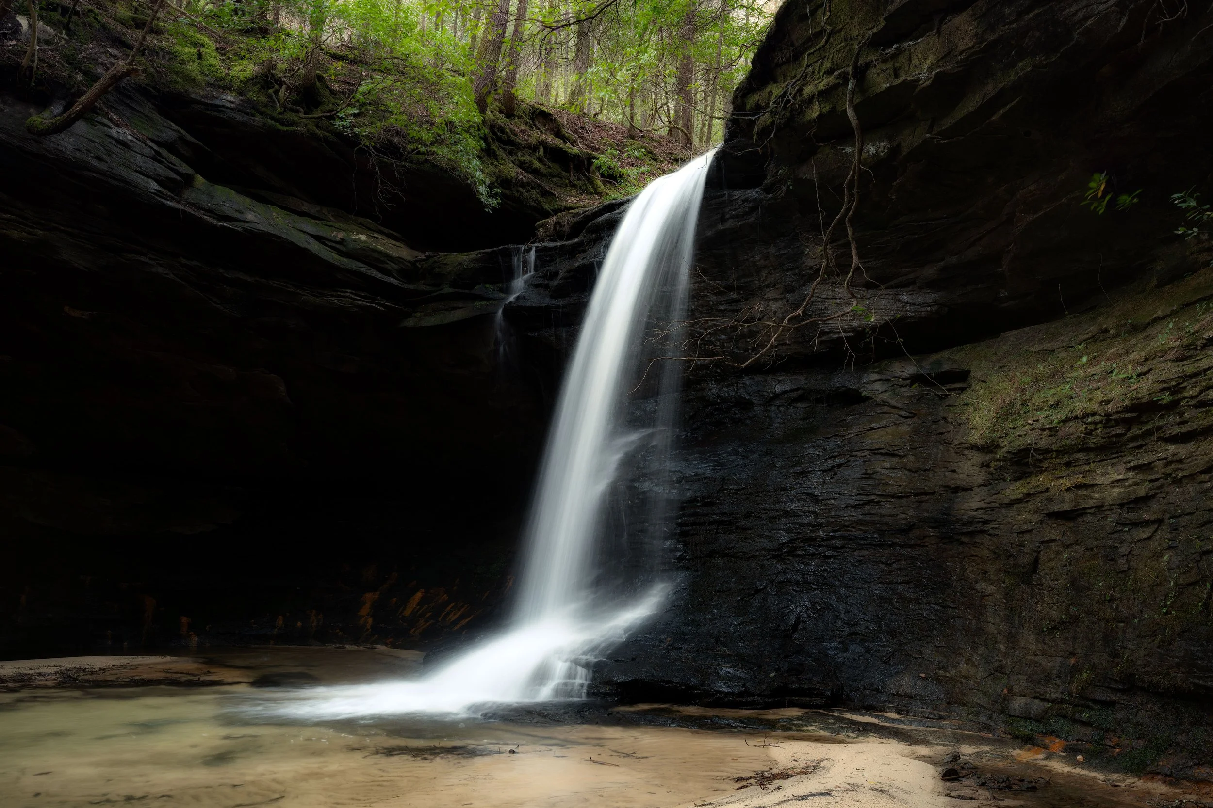 A waterfall flowing down a dark rocky cliff into a sandy pool surrounded by green trees in a forest.