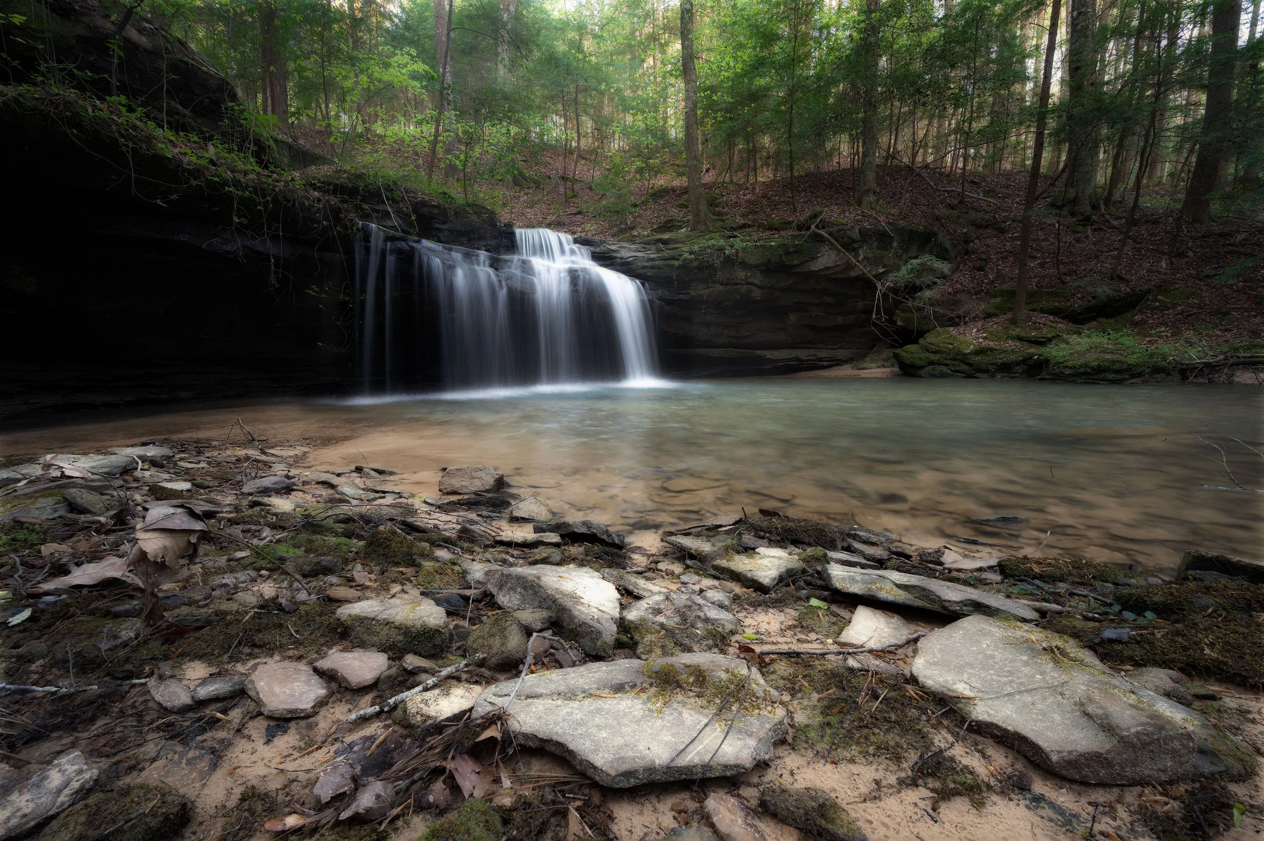 A small waterfall cascading over dark rocks into a shallow, sandy pool in a forested area with green trees and foliage.
