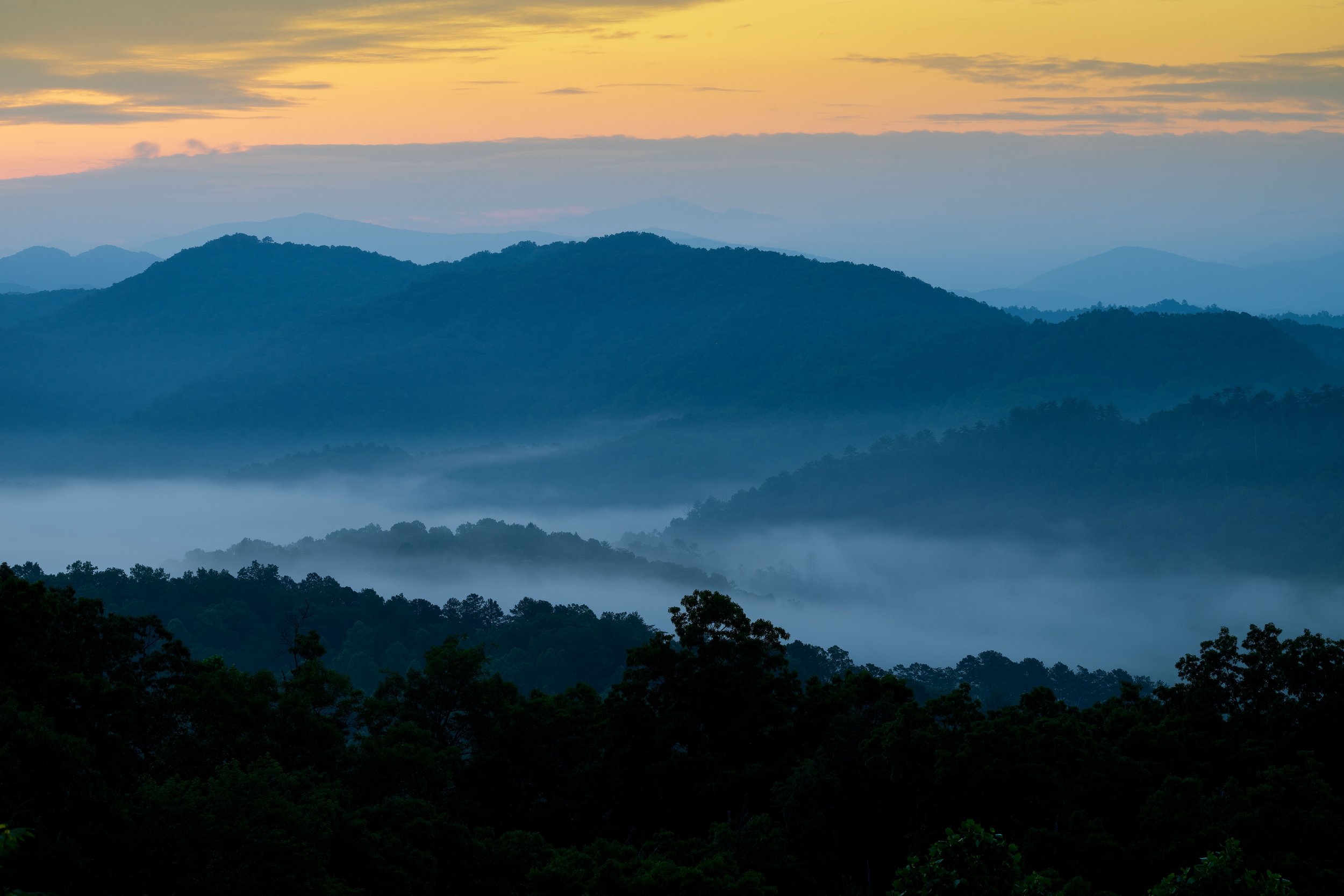 Layered mountain ranges at dawn with mist in between, colorful sky with orange and purple hues.