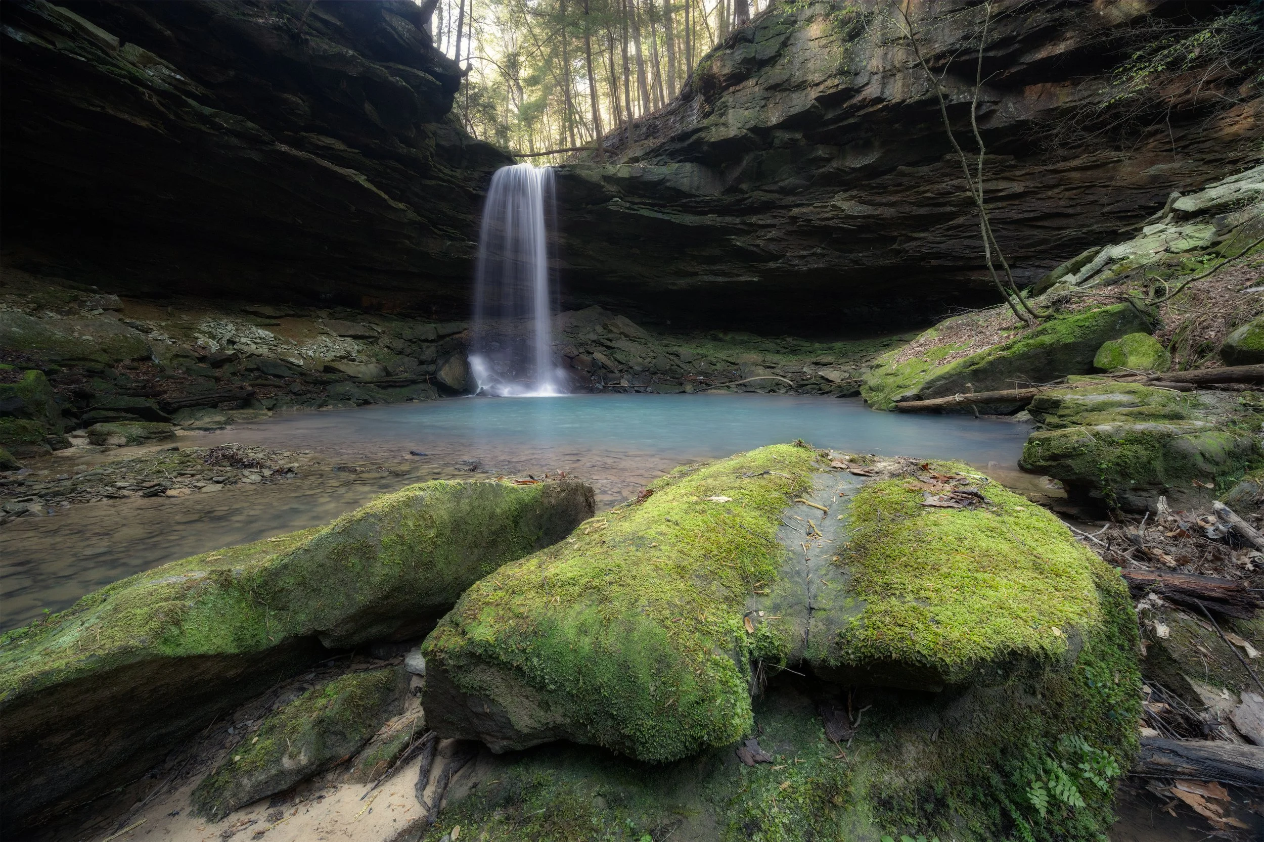 A small waterfall falls into a clear, blue pool surrounded by moss-covered rocks and dirt, with trees and shadowy rock formations overhead.