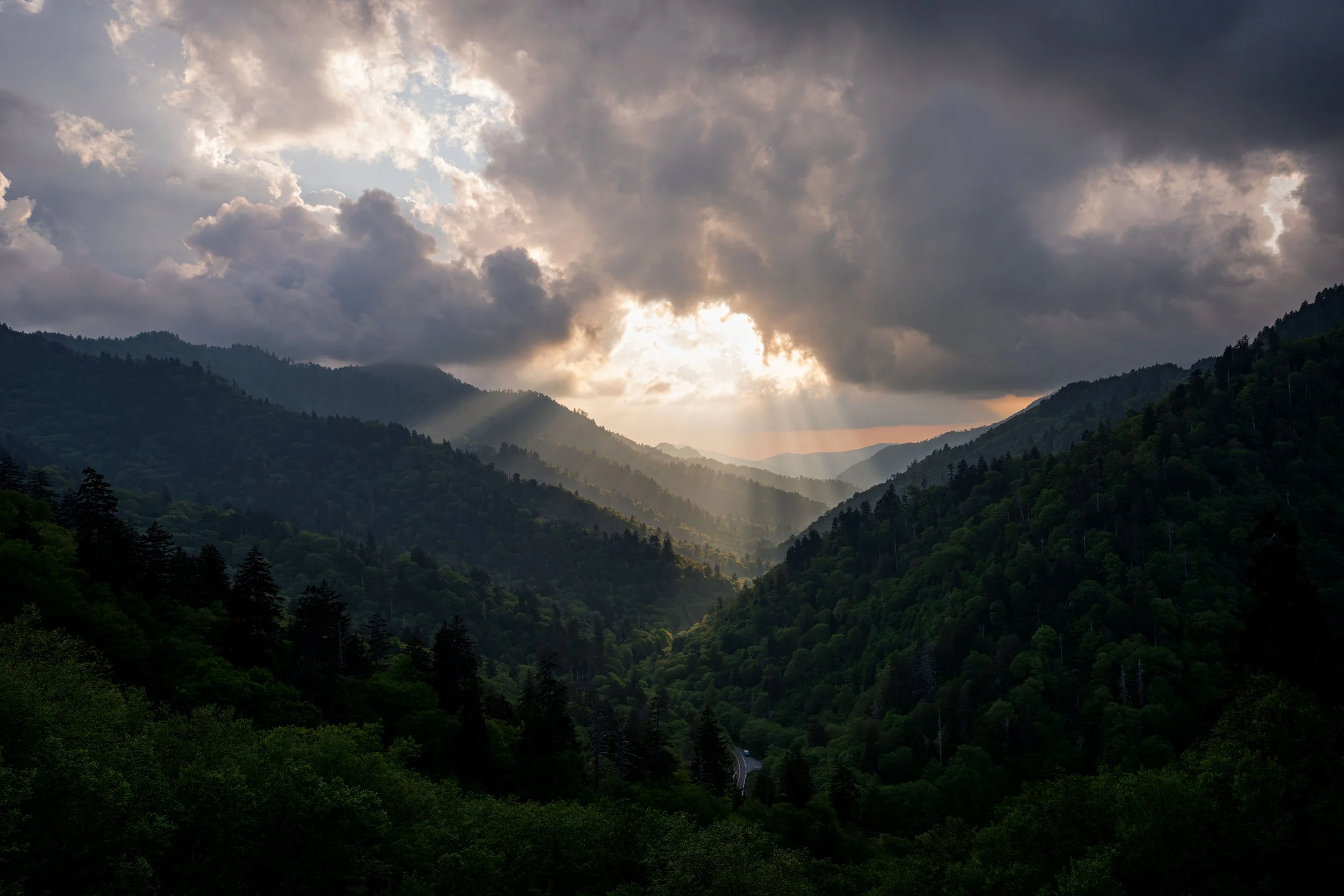 Sun rays shining through dark clouds over a mountainous forest.